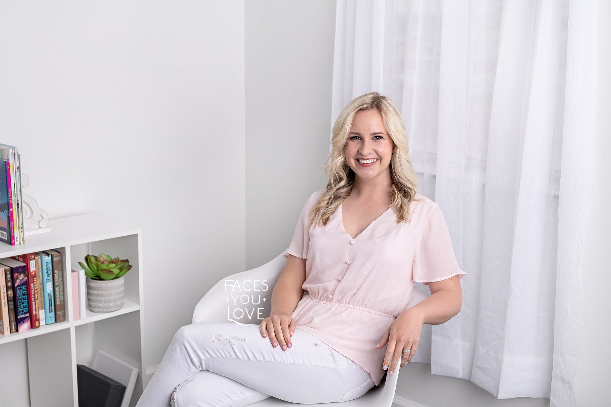 Light and airy branding photo of woman in a white chair, next to a bookshelf. She's wearing white jeans and a light pink shirt. Photographed in Kansas City by Faces You Love.