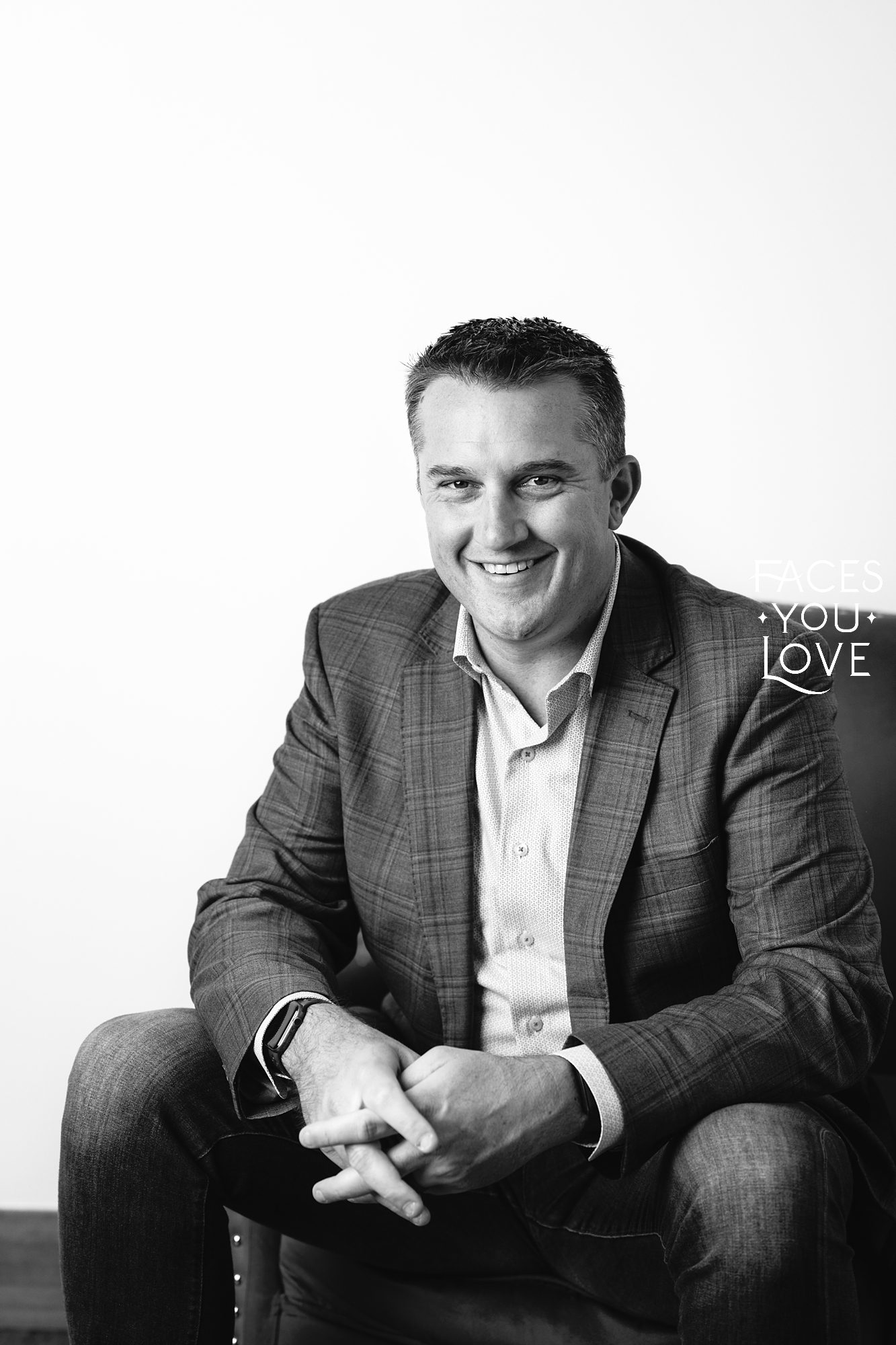 Black and white headshot of a man sitting in a chair, against a light background. He has his arms resting on his knees, in a relaxed pose, and has a smile on his face. Photographed in the Faces You Love studio in Overland Park.