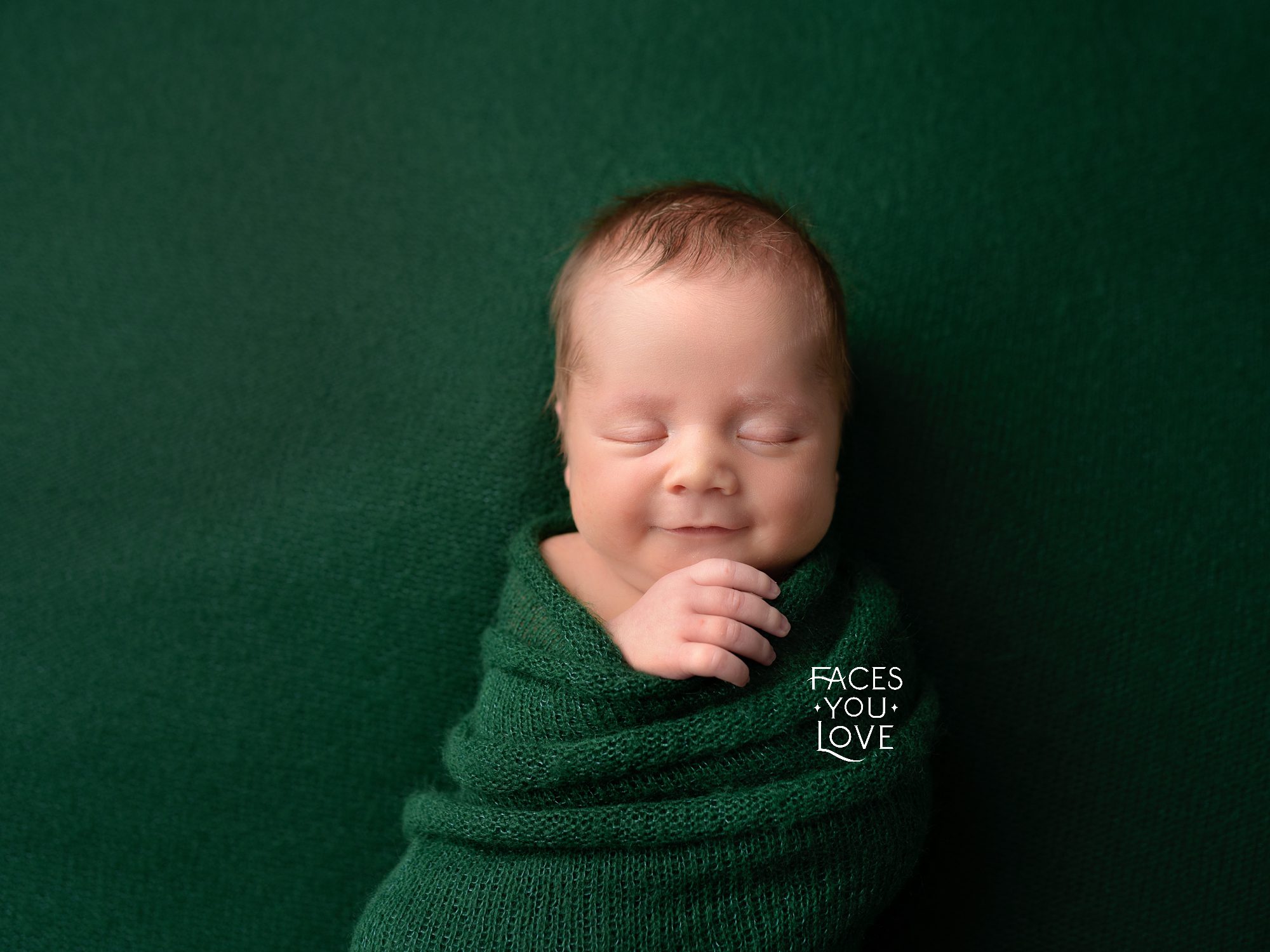 Newborn boy on forest green backdrop, and wrapped in the same color, with just one hand peeking out and resting under his chin. Baby has a slight smile on his face. Picture done by Overland Park baby photographer Helen Ransom of Faces You Love.
