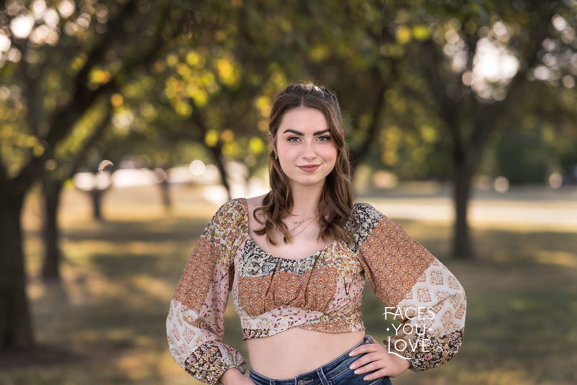 High school senior girl, standing in a grove of trees, with her hands on her hips. She's wearing jeans and a patterned crop top, and has a soft, closed mouth smile. Photographed in Overland Park by Faces You Love Photography.