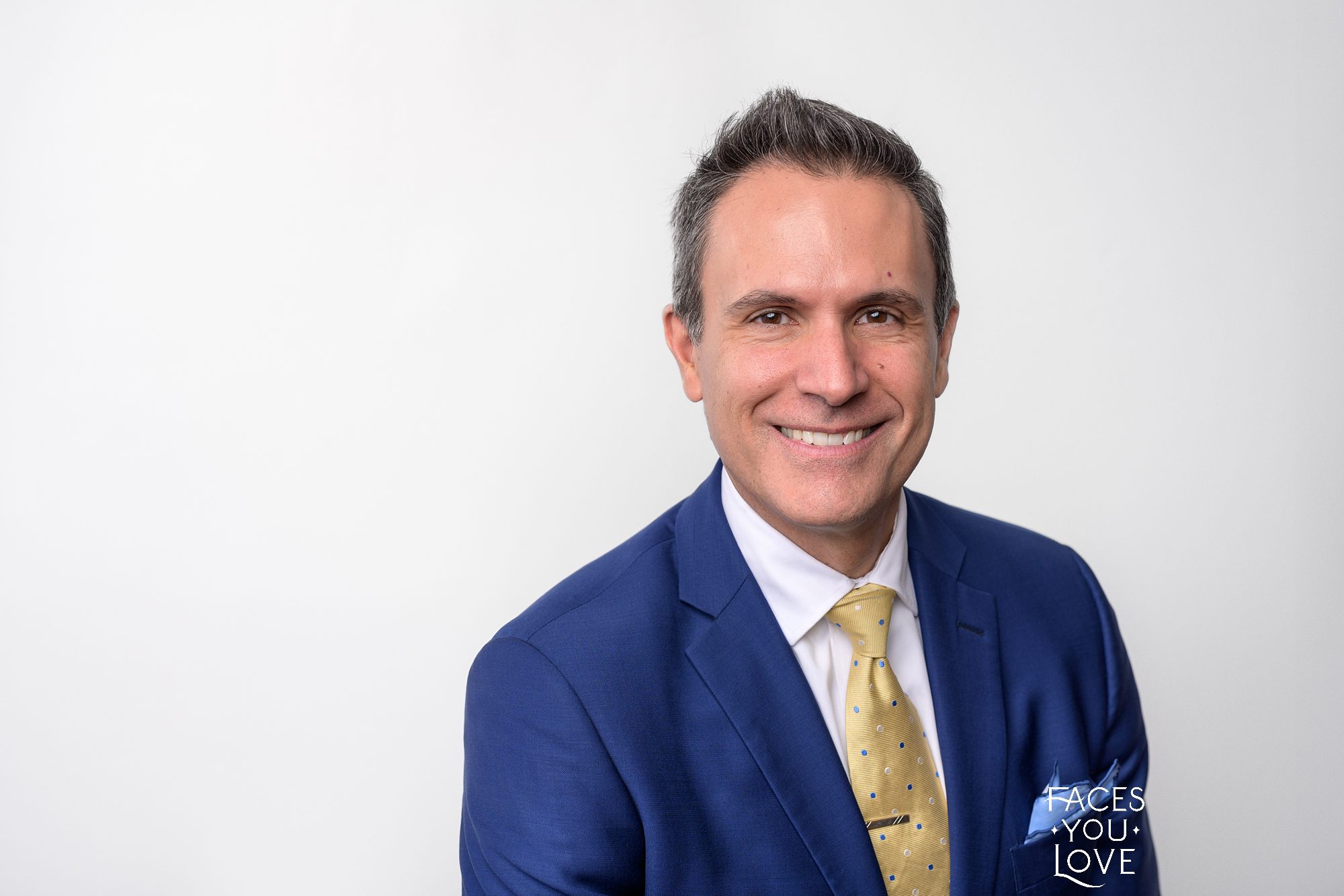 Closeup male headshot photographed in Kansas City at Faces You Love Photography. Man is smiling and wearing a navy blue suit and yellow tie, against a white background.