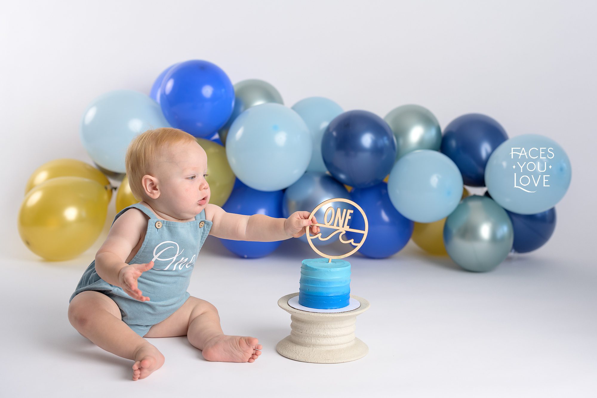 One year baby session, photographed on a light gray background. Baby is wearing a blue onesie with light brown buttons. Photographed at Faces You Love Studio in Overland Park.