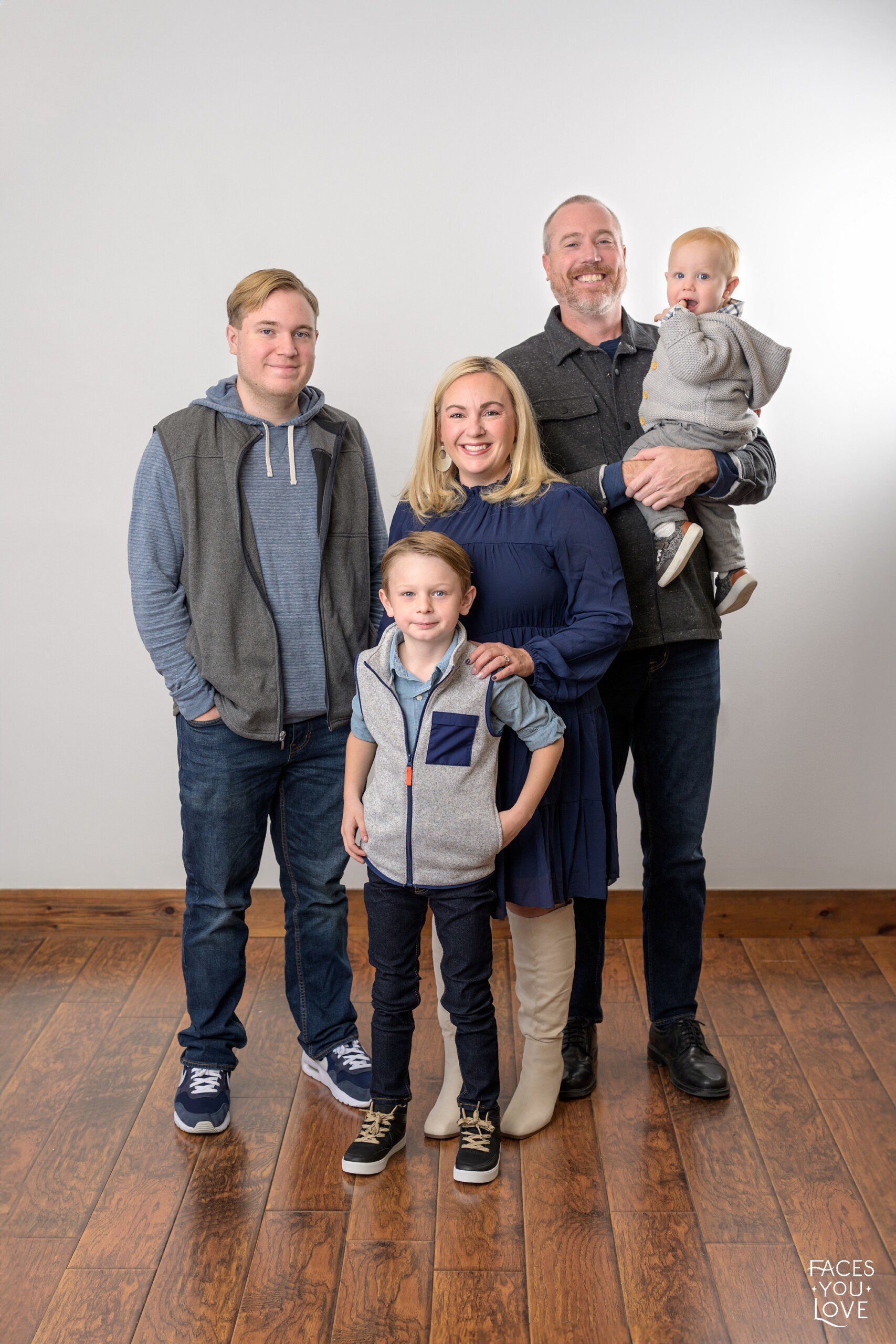 Family watch me grow session, photographed on a gray background. They are wearing shades of gray and blue. Photographed in studio at Faces You Love in Overland Park.