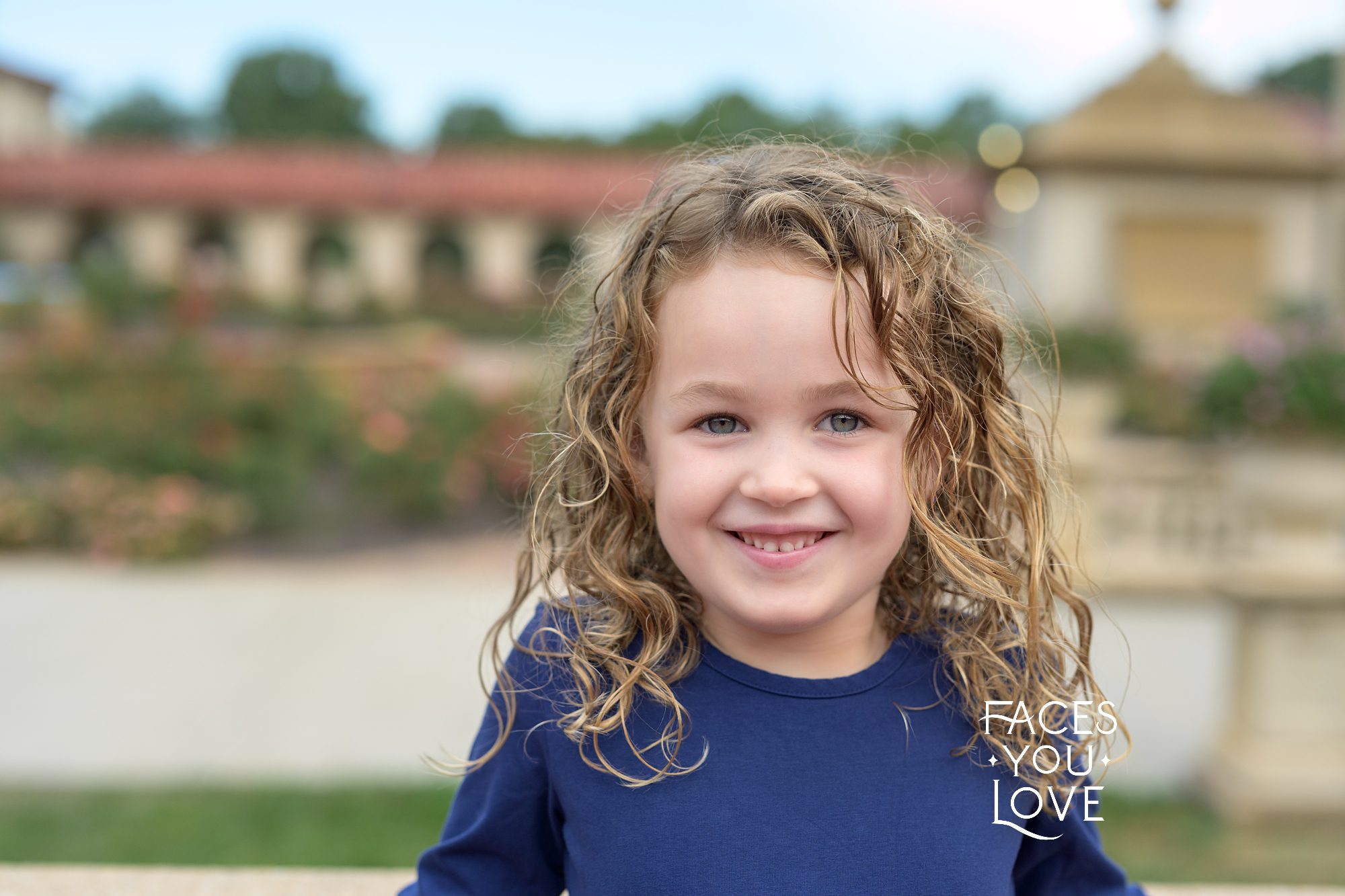 Close up of a young girl, with curly hair and blue eyes. She's standing overlooking the rose garden at Unity Village. Photographed by Helen Ransom of Faces You Love, located in Overland Park, KS.