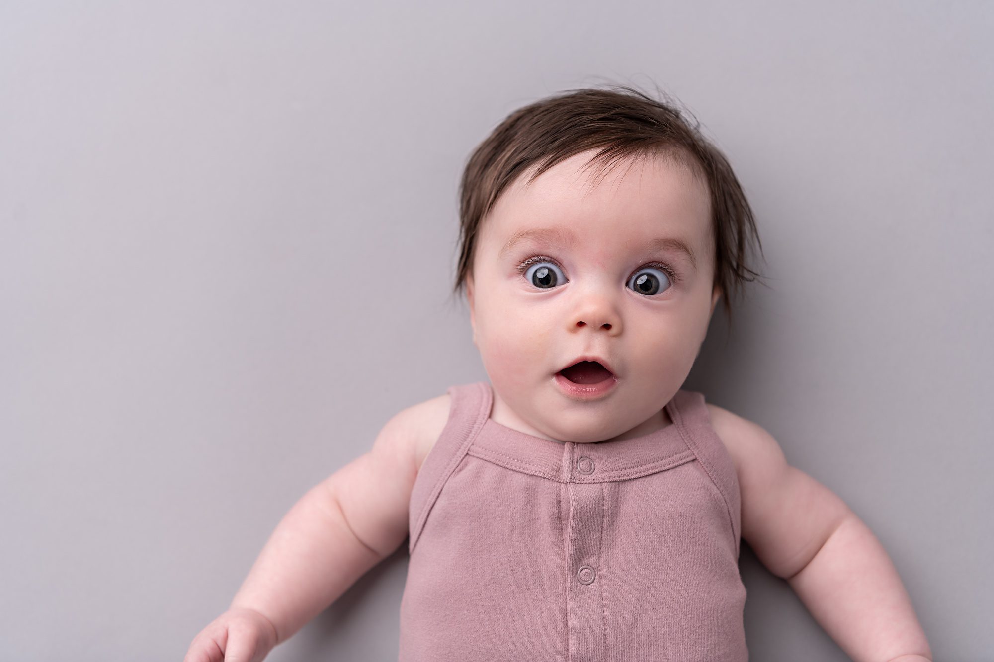 Wide-eyed baby girl with dark hair, laying on a light mauve backdrop at a photo studio in Kansas City.