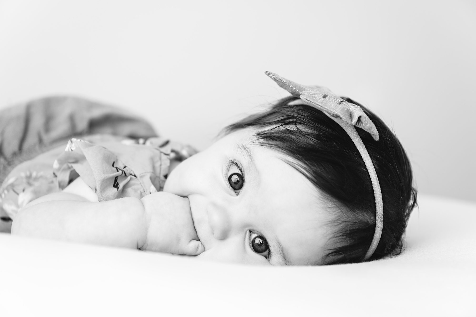 Black and white photograph of a baby girl laying on her stomach, with her hand in her mouth. She's making eye contact with the camera and best baby photographer Helen Ransom.