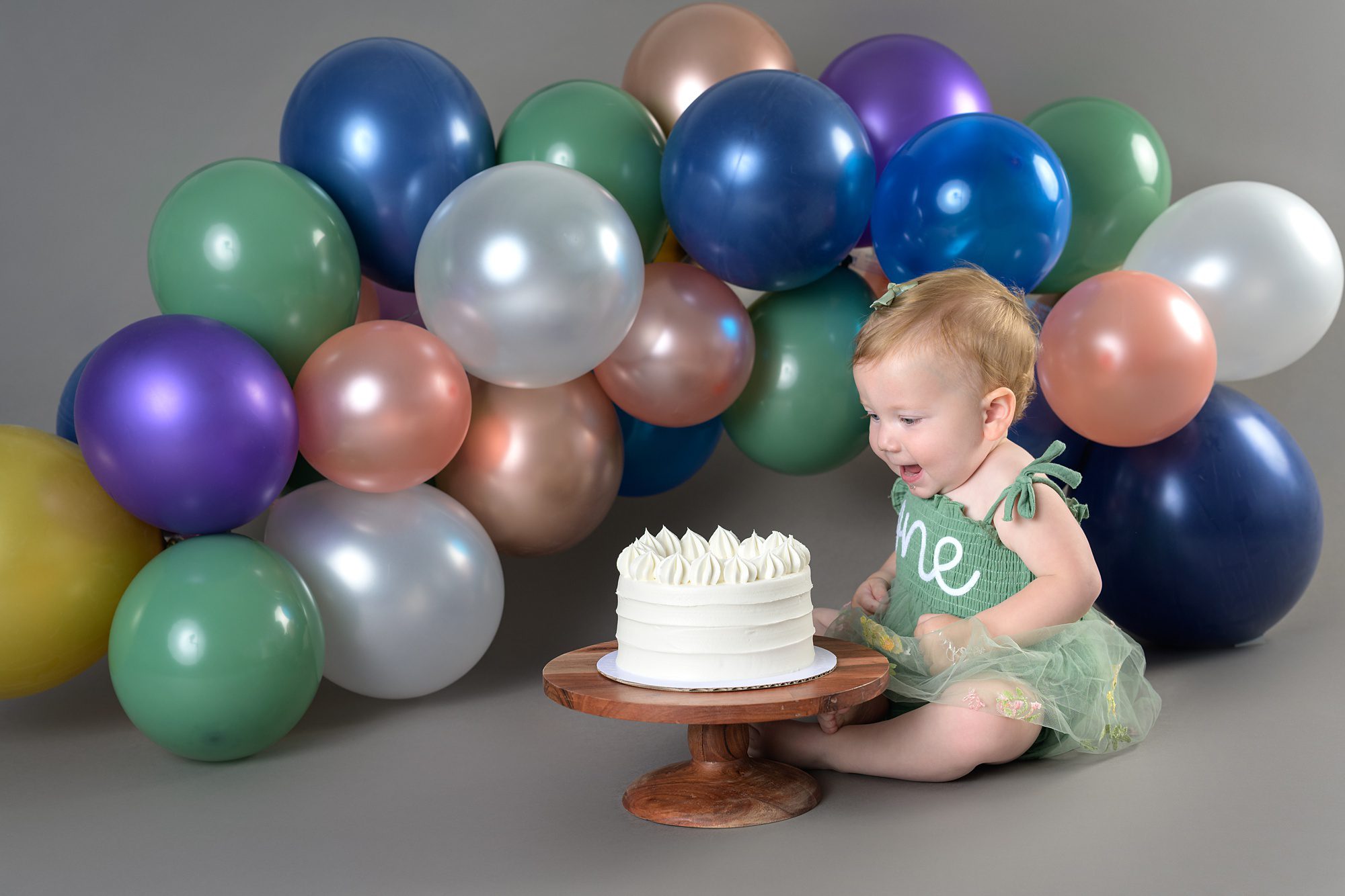 Baby girl smiling at the cake sitting in front of her, at her cake smash photography session. She's on a gray background and has a balloon arch behind her made up of blues, greens, and peach.