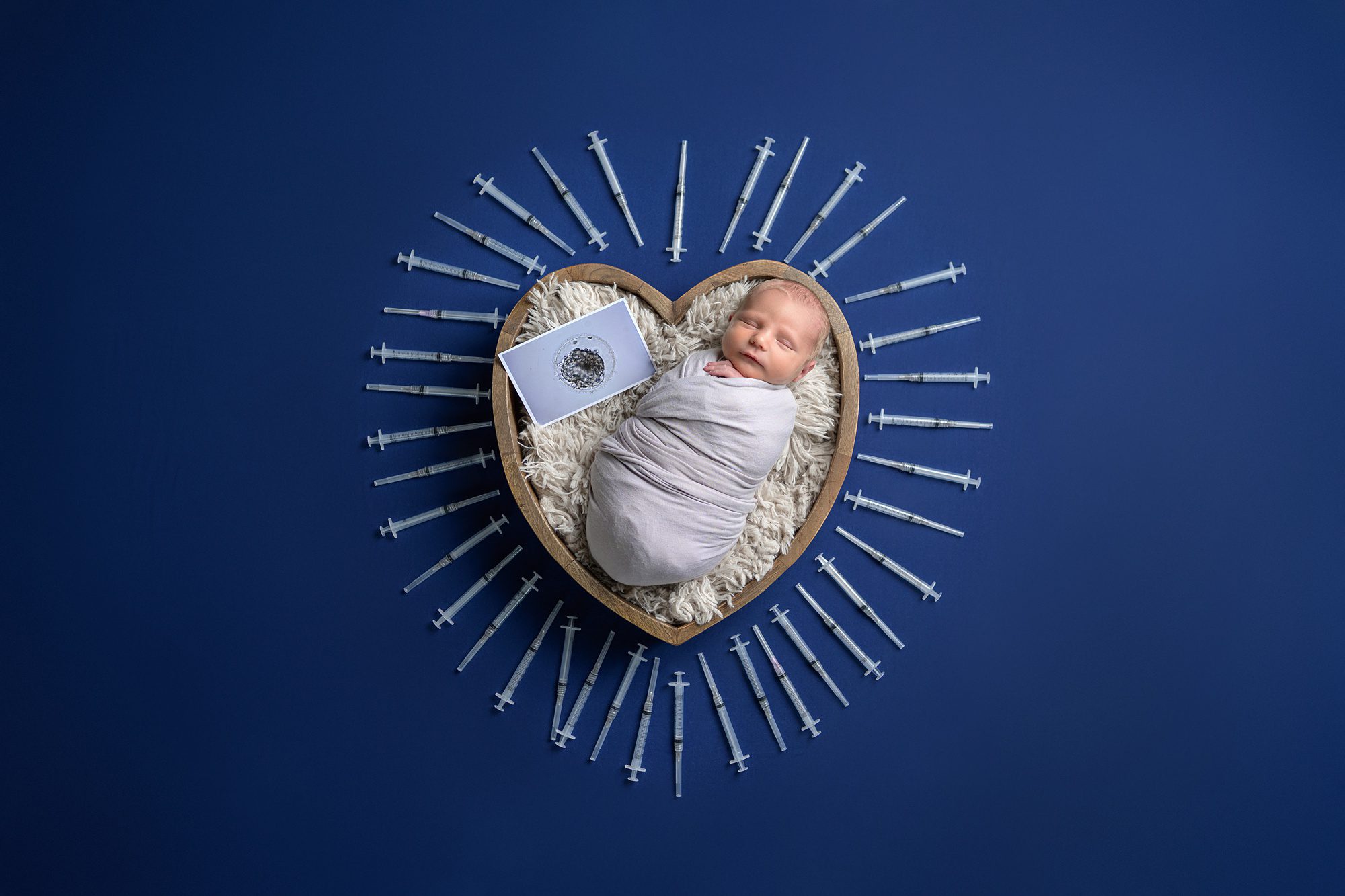 Newborn photo that is a tribute to the parents IVF journey. Baby is in a heart shaped bowl with an embryo picture, and the bowl is surrounded by empty IVF syringes. Photographed by one of the best newborn photographers in Kansas City, Helen Ransom.