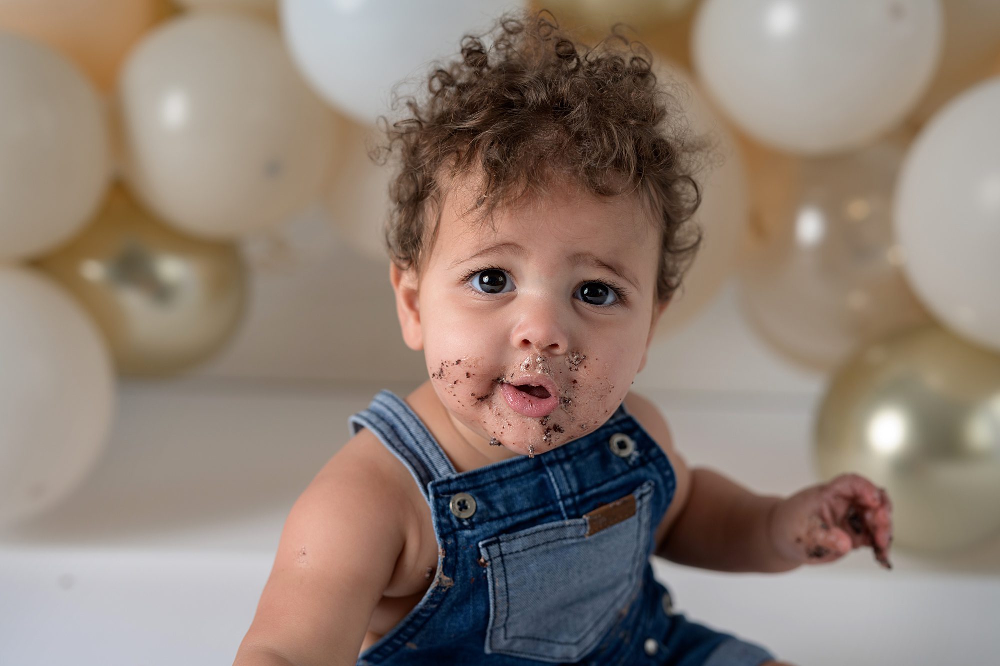 Close up of a baby mid cake smash photoshoot. They're wearing overalls, have dark, curly hair, and chocolate cake and frosting all over their face.