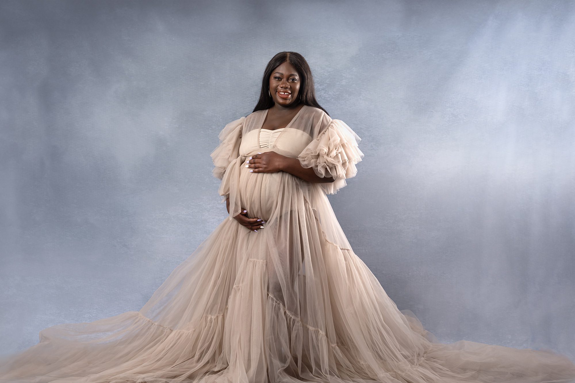 Pregnant African American woman, standing against a blue studio backdrop and wearing a light brown robe made of tulle, provided by the Kansas City maternity photo studio.