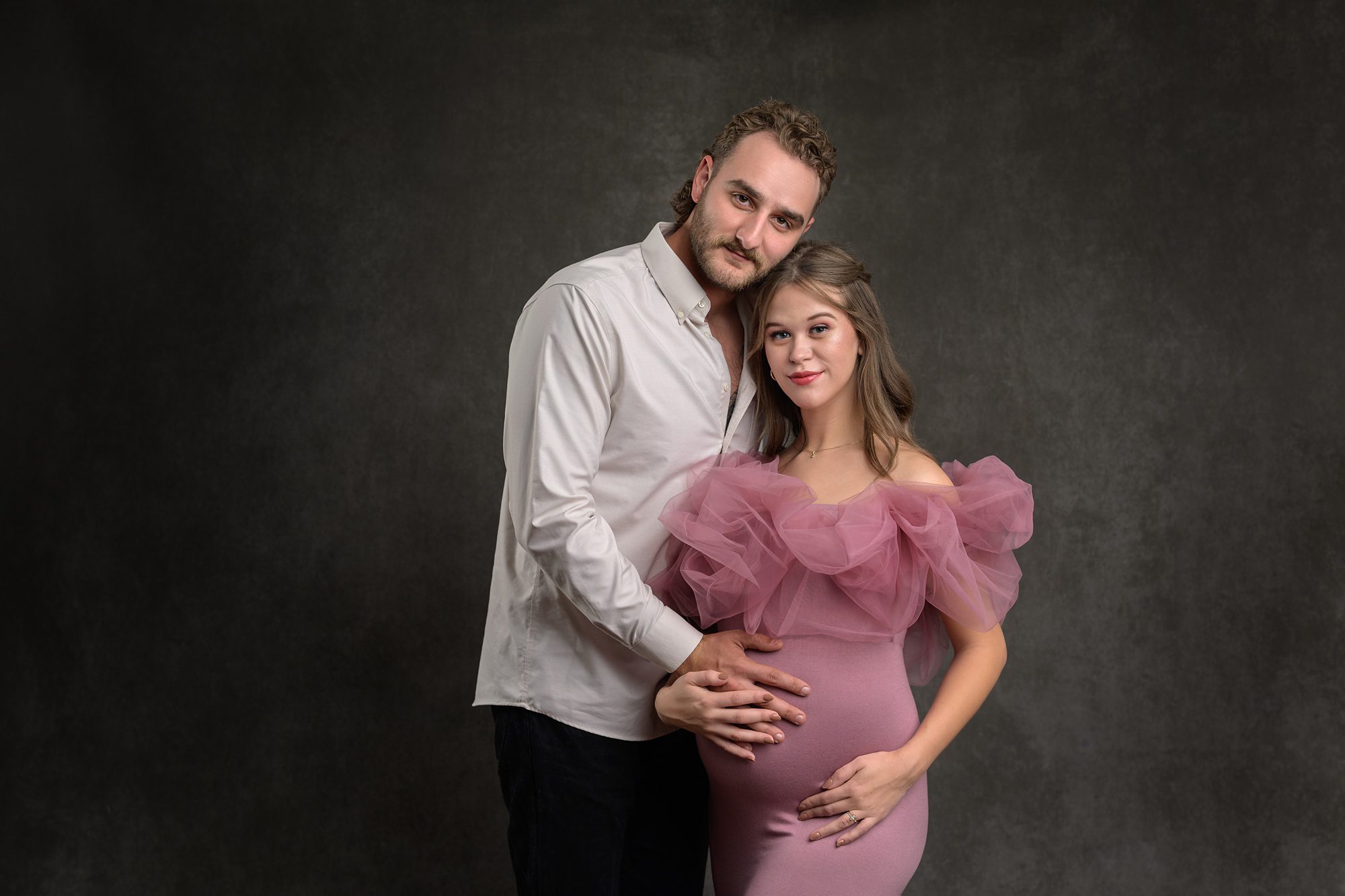 Couple posing against a dark background at a pregnancy photo session. Dad is wearing a white button down and mom is in a pink gown with tulle on top.