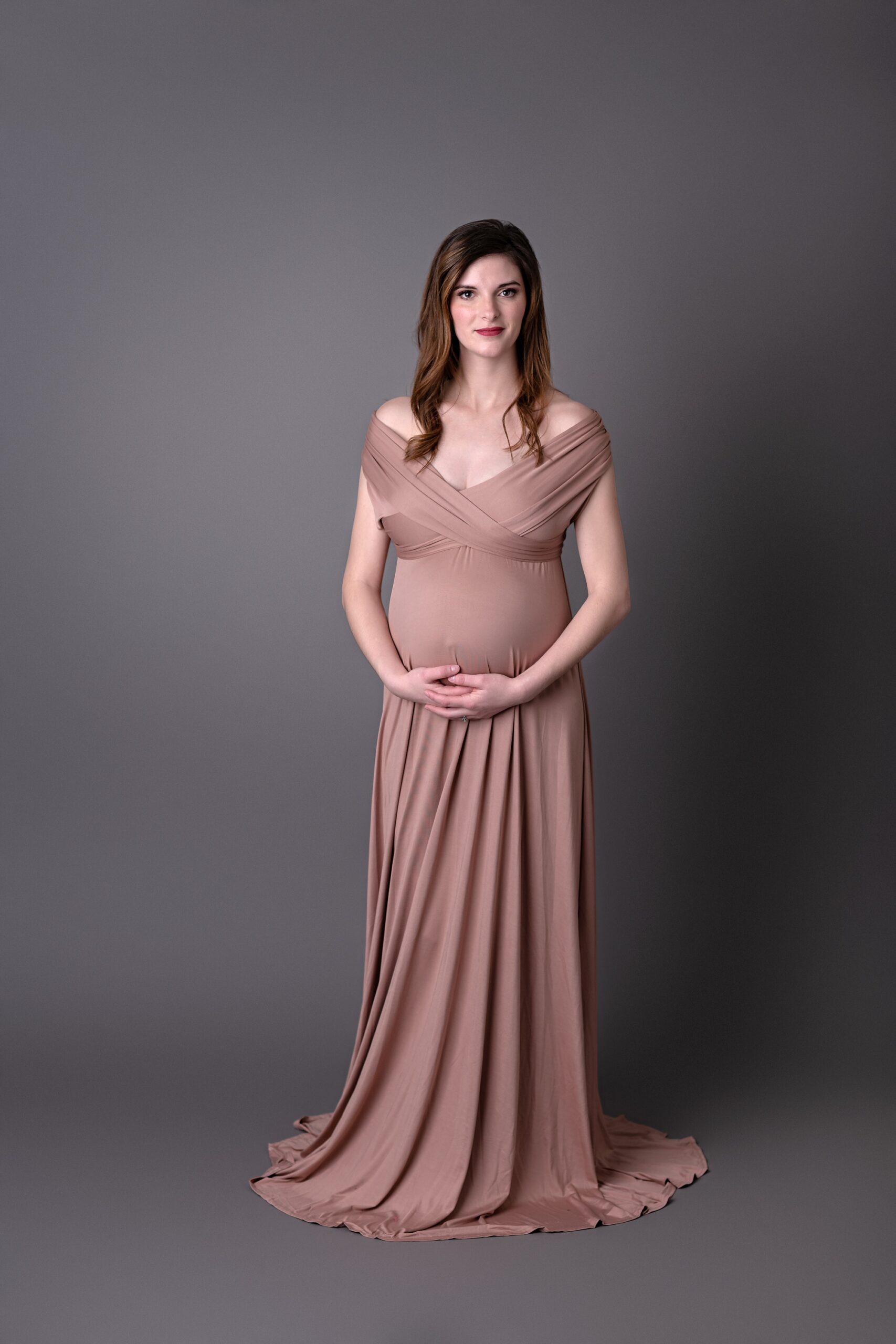 Pregnant woman in a rose gold dress, standing on a gray background by Kansas City photographer Helen Ransom.