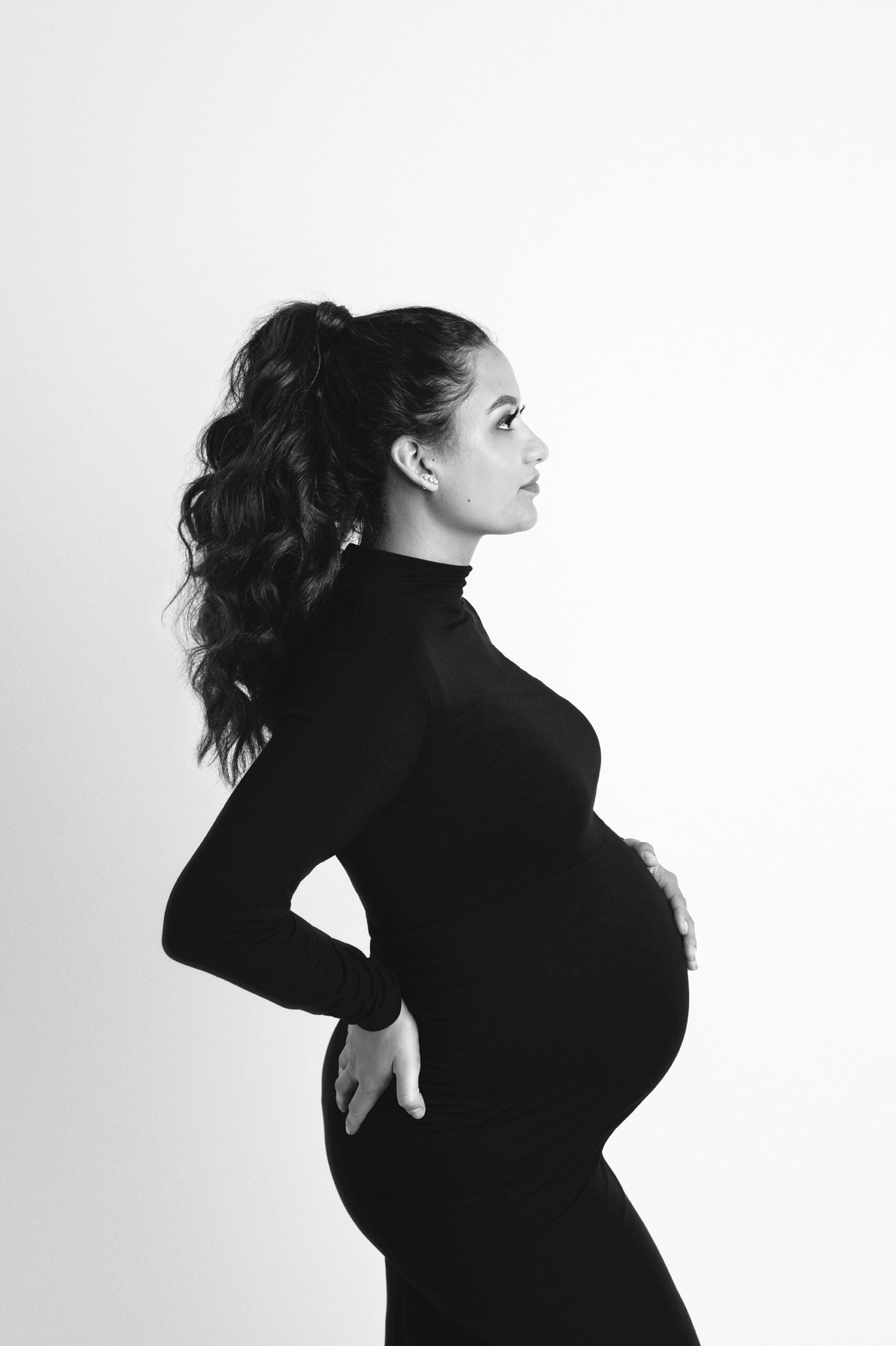 Simple maternity portrait in black and white. Woman is facing to the side, wearing a sleek form fitting gown with hair in a high ponytail. Photographed at a maternity photo studio in the Kansas City metro.