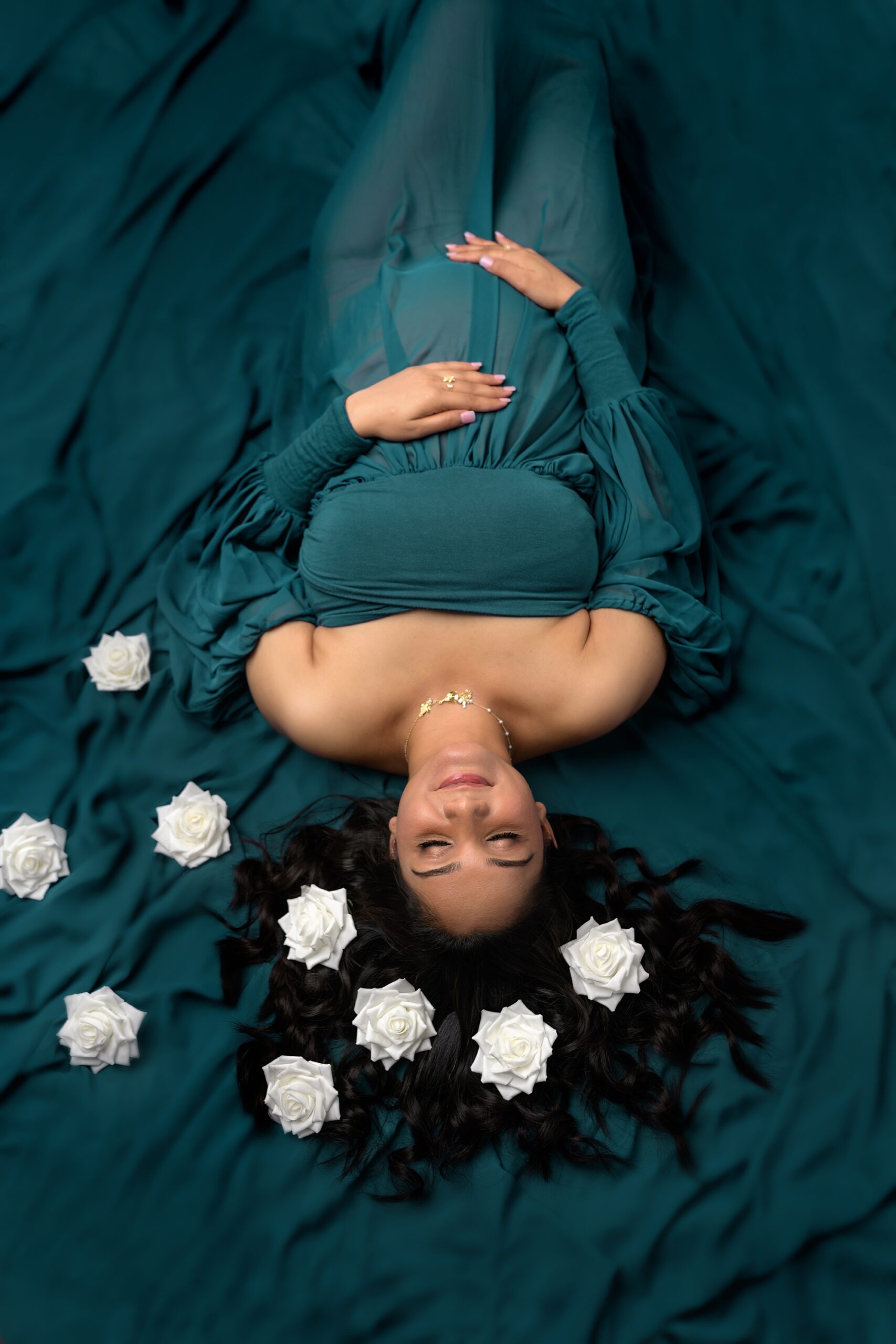 Pregnant mom laying on her back, with hair splayed out, white roses scattered around her head, and a serene look on her face. Photographed by maternity photographer Helen Ransom