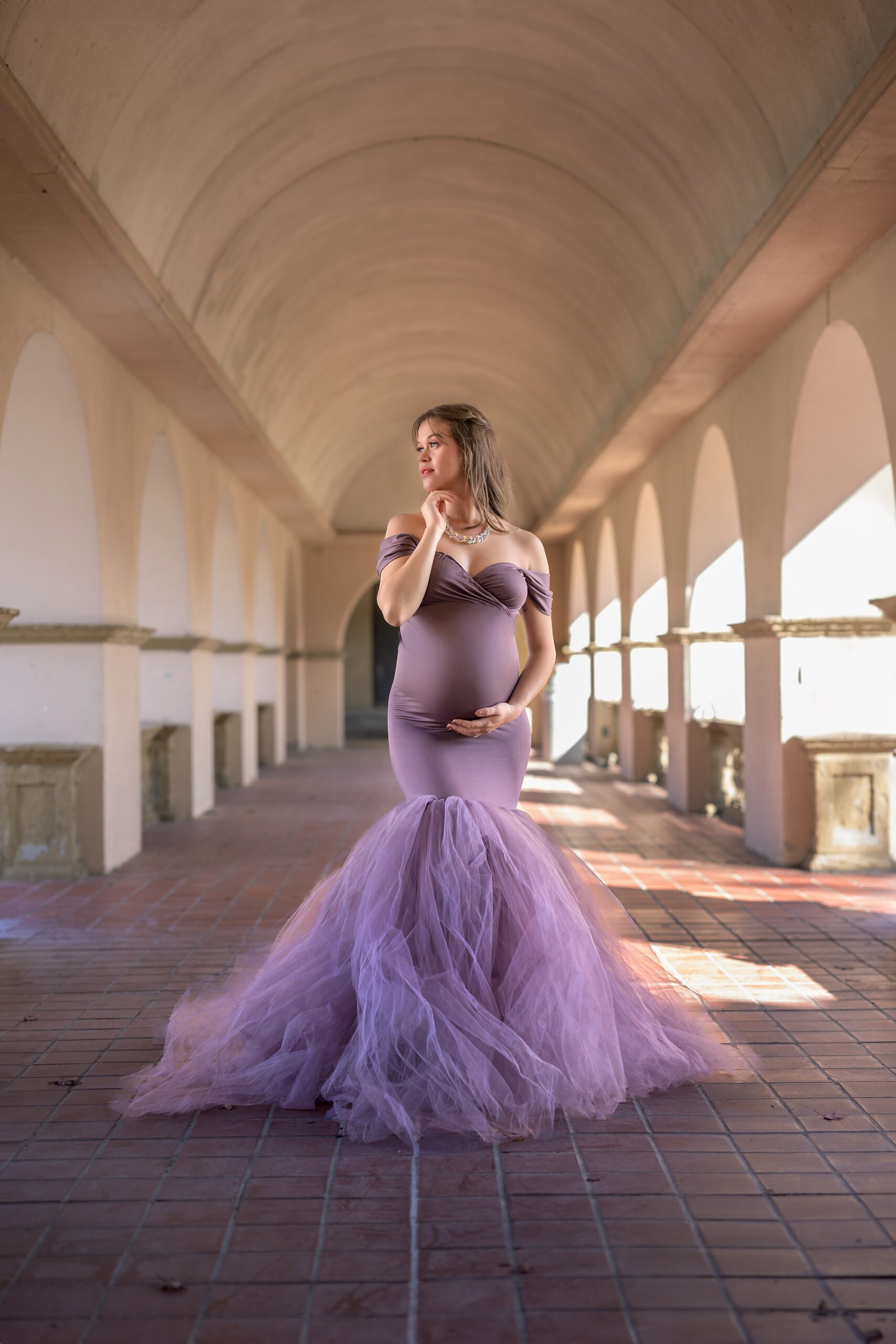 Glamorous maternity photograph made at Unity Village in the Kansas City area. Mom to be is in a mermaid style purple gown, standing in a row of arches, and gazing to the left.