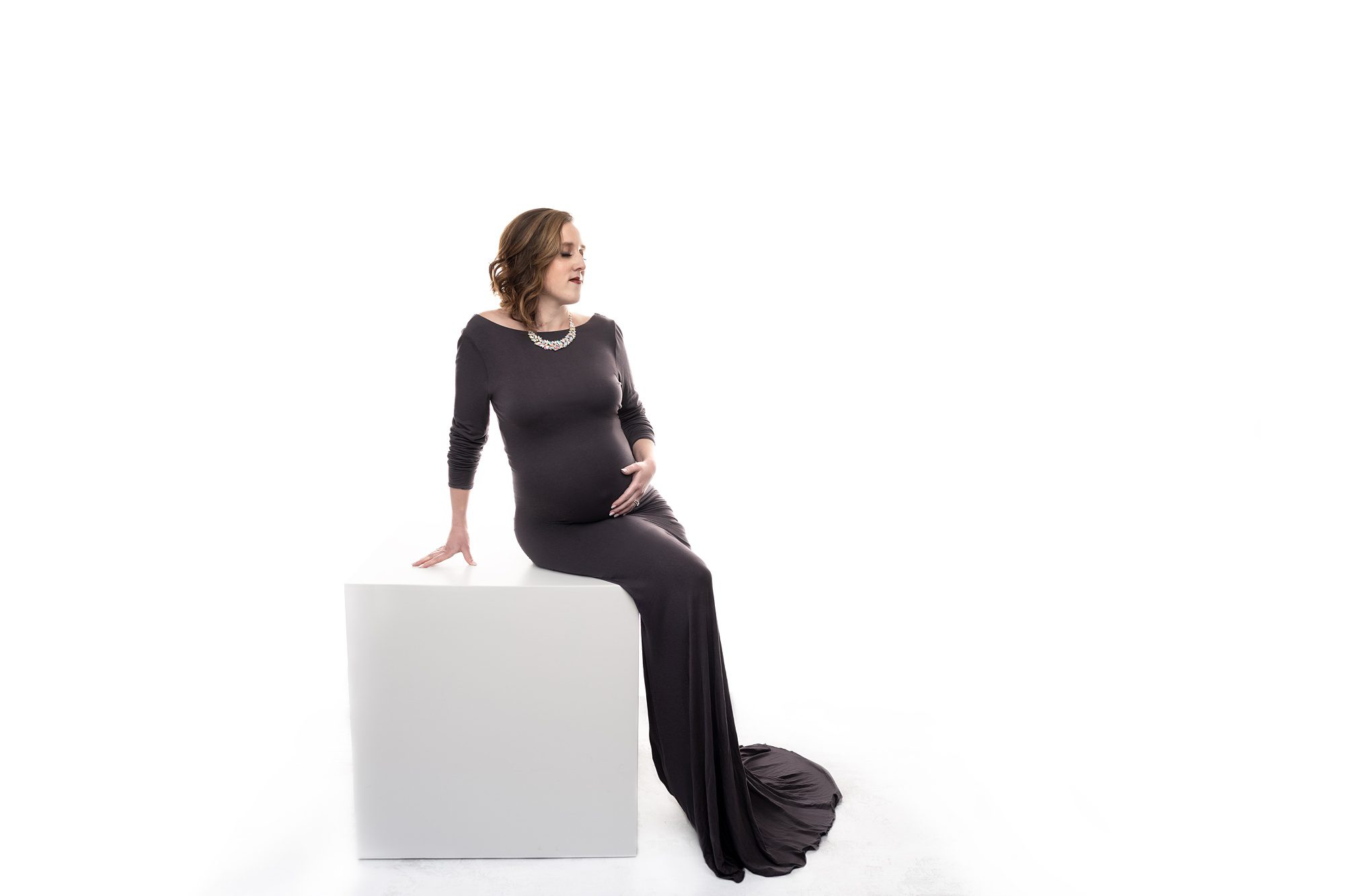 Elegant but simple maternity photo, of pregnant mom in a dark gray, form fitting, floor-length gown, in a completely white set, sitting on a white wooden box.
