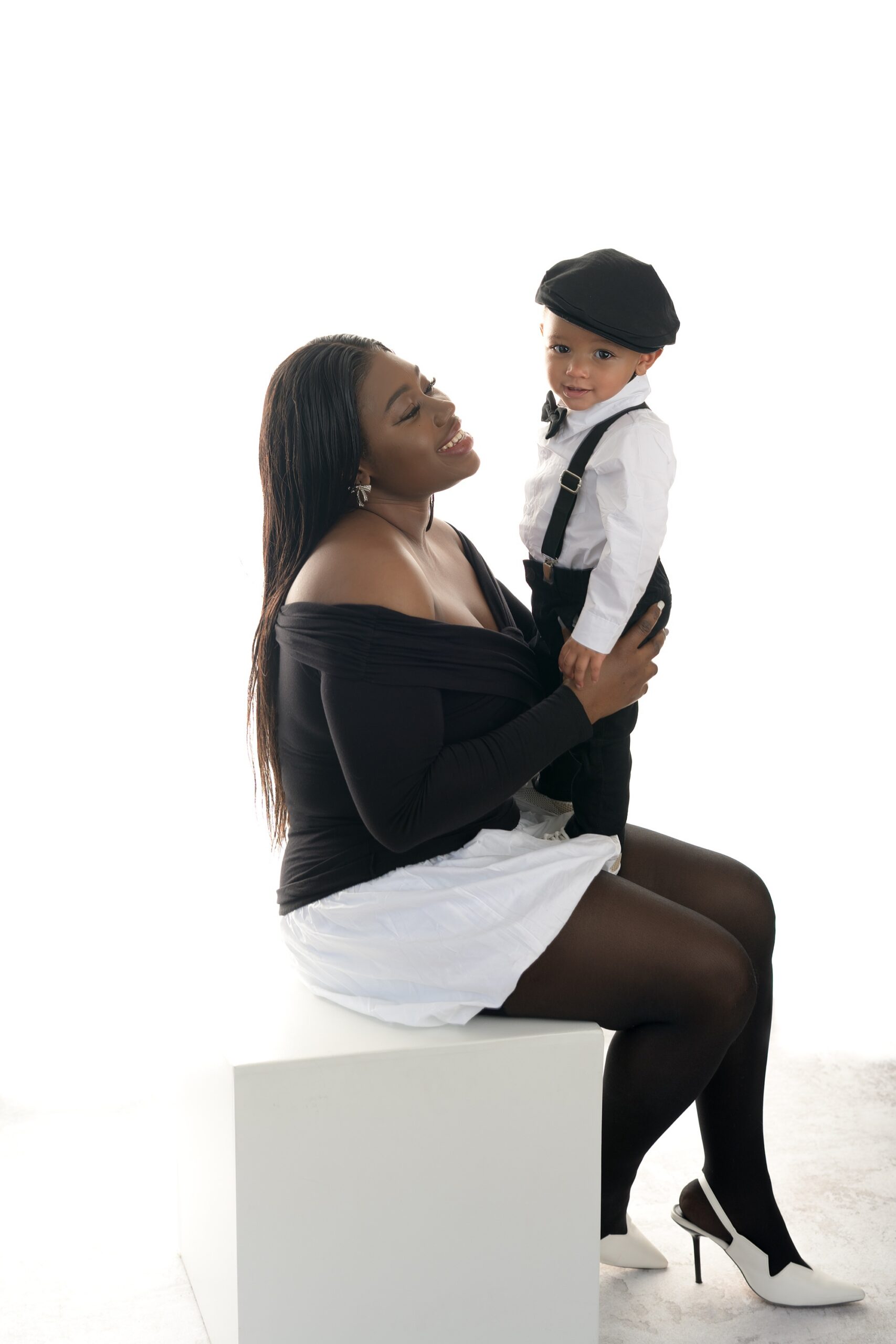African American mom and her baby at a mommy and me photoshoot. They're on a white background, wearing all black and white themselves. Mom is sitting on a white cube and the baby is standing in her lap.