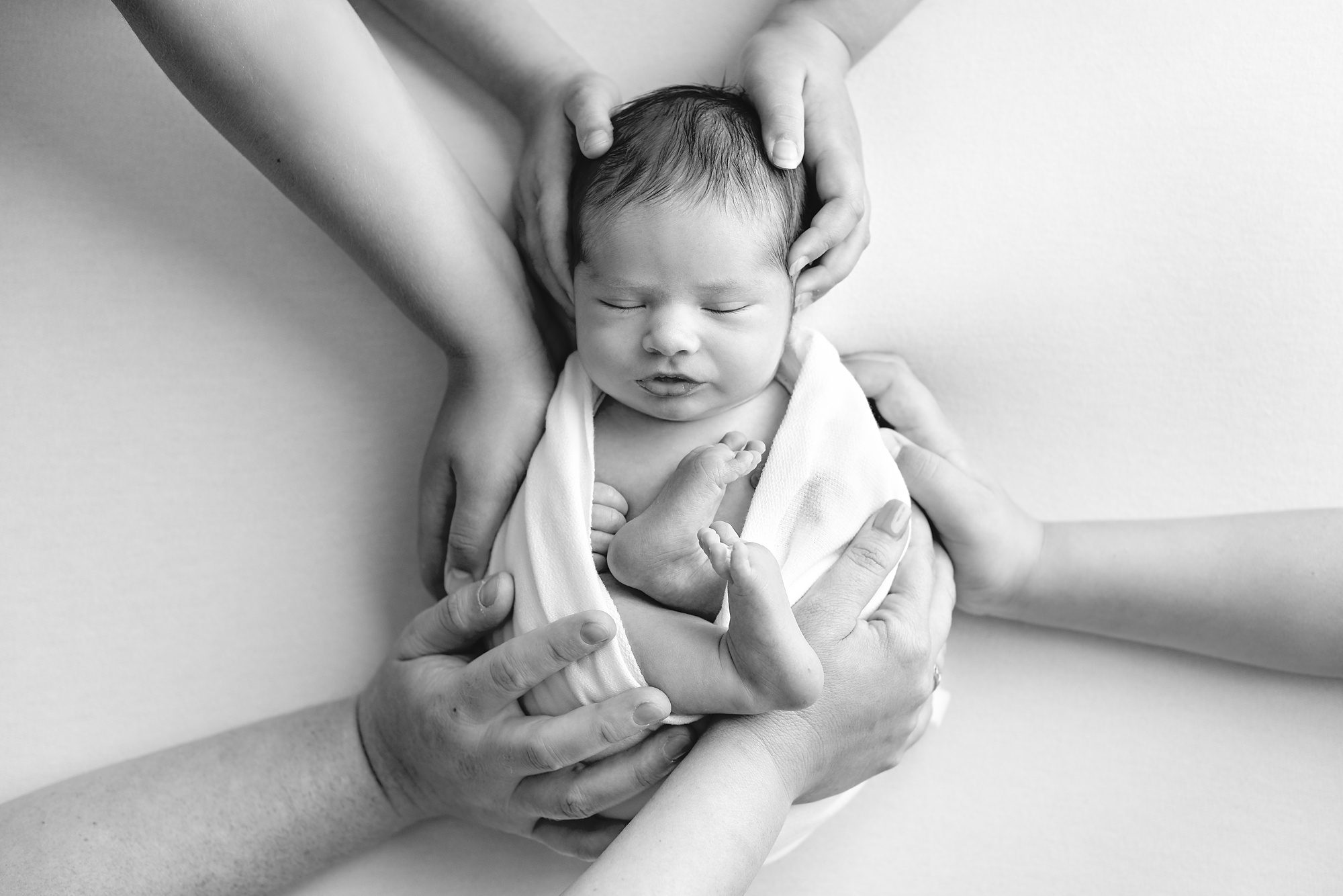Black and white image of a newborn baby surround by the hands of his parents and four older siblings, taken at his new born photoshoot by Helen Ransom.