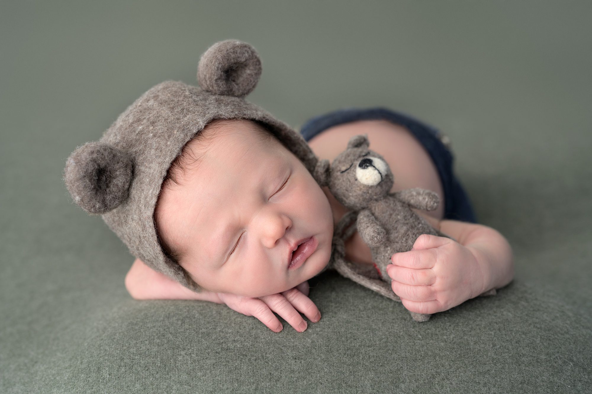 Newborn baby on a sage green backdrop, laying on his belly with one hand under his head and the other clutching a tiny teddybear, and baby is wearing a wool bonnet with bear ears on it.