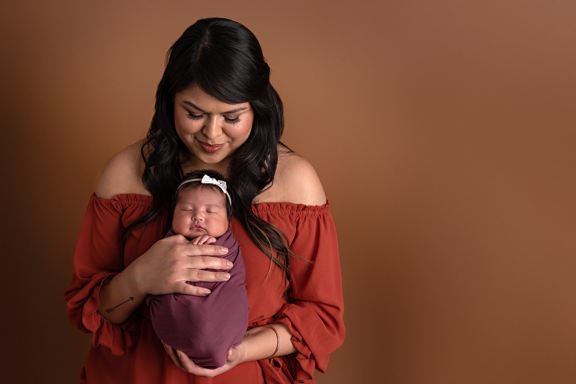 Native American mom and newborn baby, standing against a copper colored studio backdrop at their Kansas City newborn photo session. Baby is wrapped in purple and being held against mom's chest, and mom is wearing a rust colored off the shoulder top and looking down at her baby.
