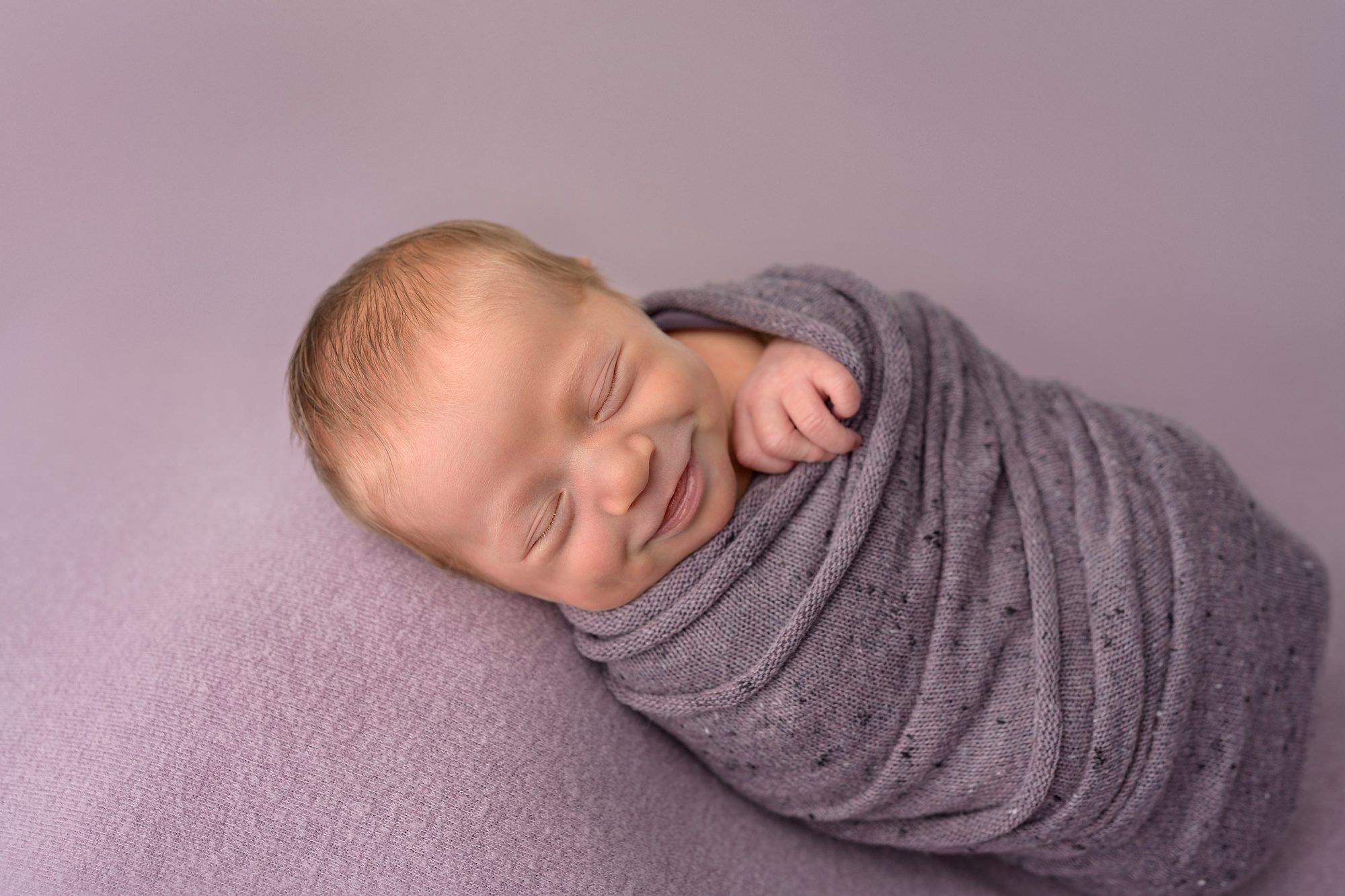 Newborn baby girl with a big smile, while she is wrapped in purple and laying on a purple backdrop at a newborn photography Kansas City studio run by Helen Ransom.