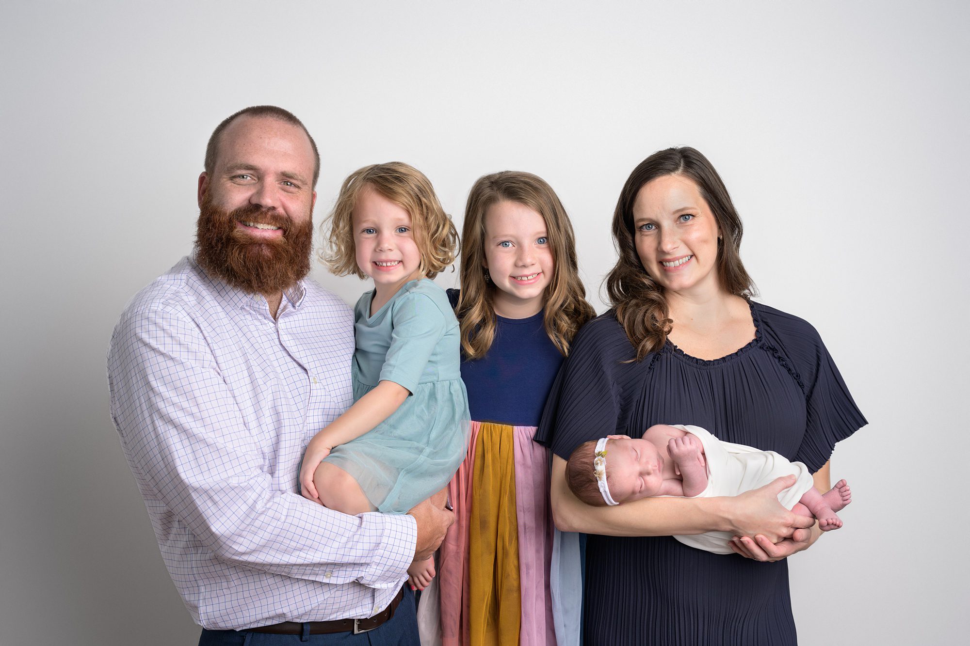 Newborn and Family at a photography session. Family is standing against a while wall, dad is holding the middle sister, oldest sister is standing in the middle, and then mom is holding the newborn baby.