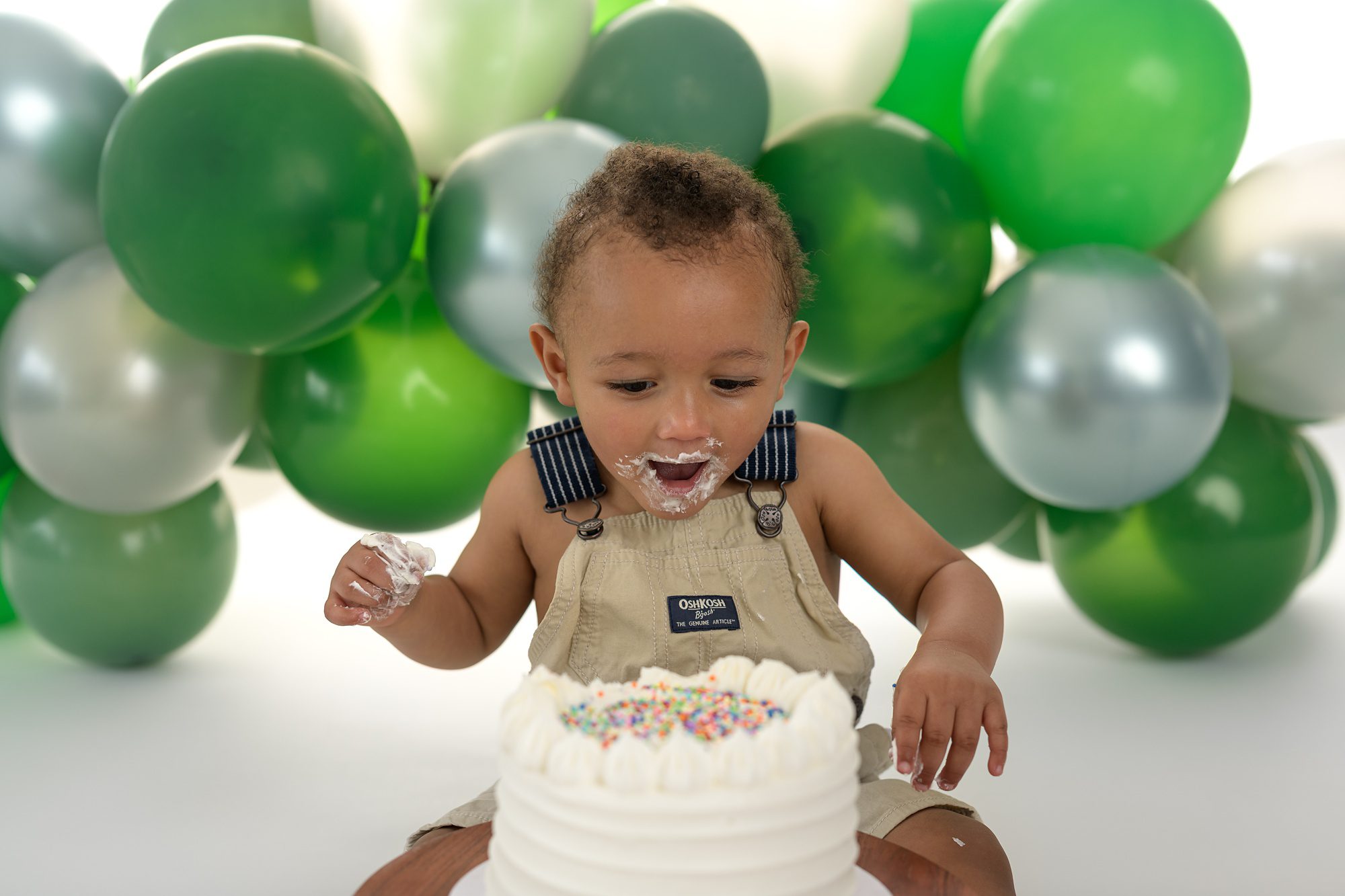 Baby book excited to see his cake at his one year photo session in Overland Park. Cake is white with colored sprinkles on top, and behind him is a balloon arch of green and silver balloons.