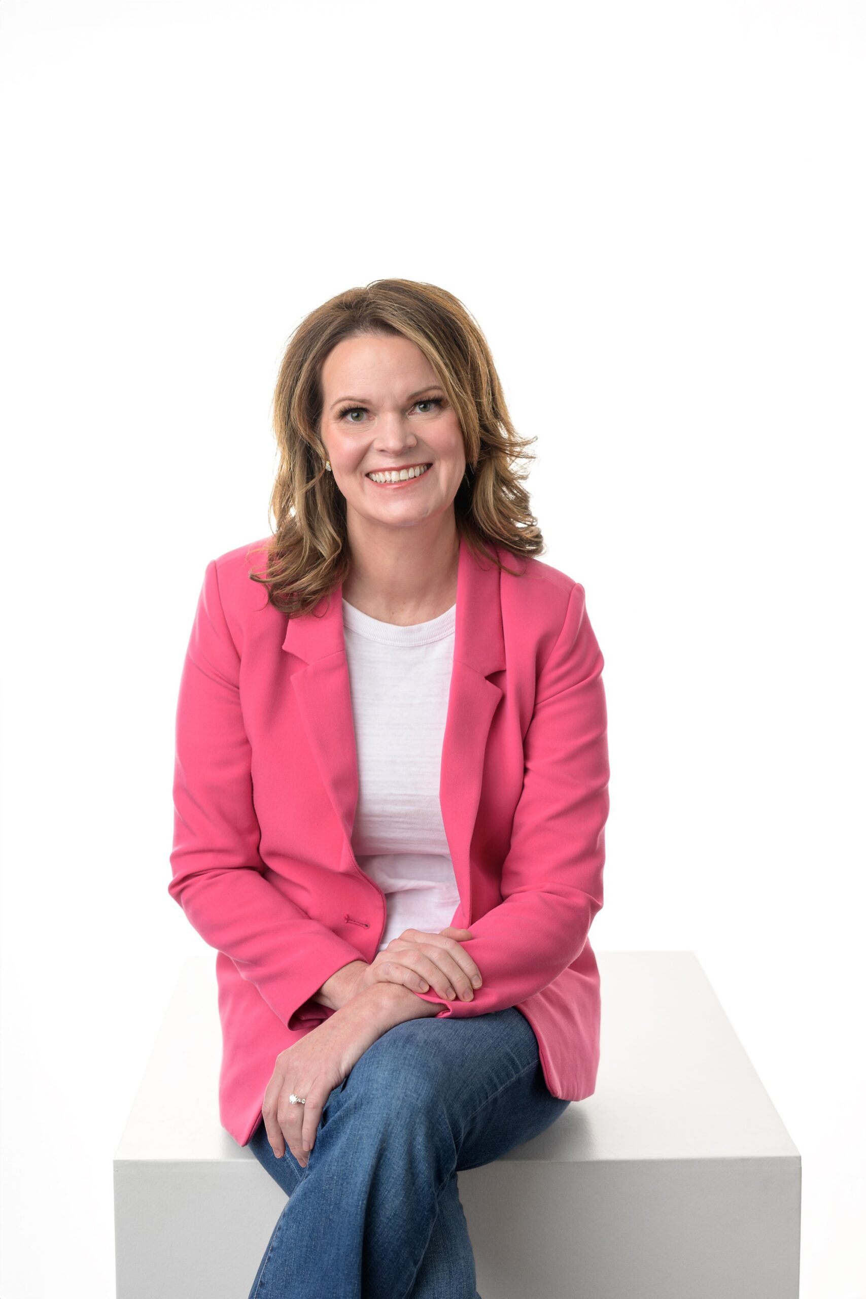 Woman in jeans, white top, and pink blazer sitting on a large white box against a bright white background, as part of her branding shoot portfolio with Helen Ransom Photography.