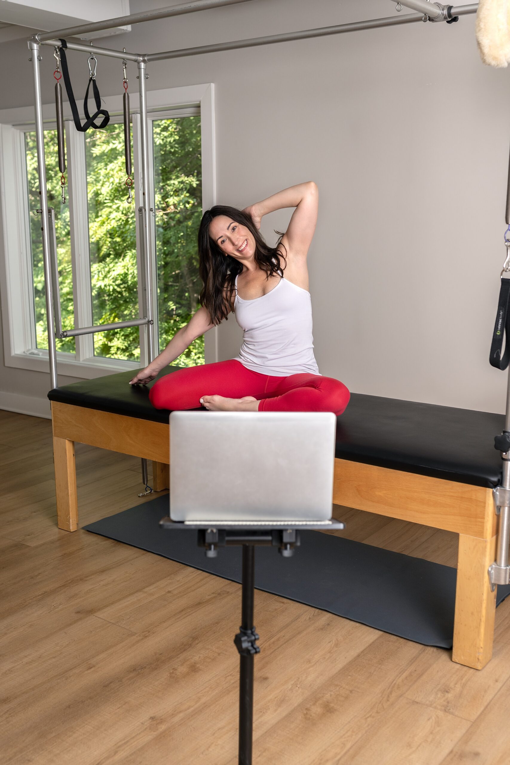 Fitness instructor in a white tank and red leggings stretching in front of a lap top while leading a virtual fitness class.