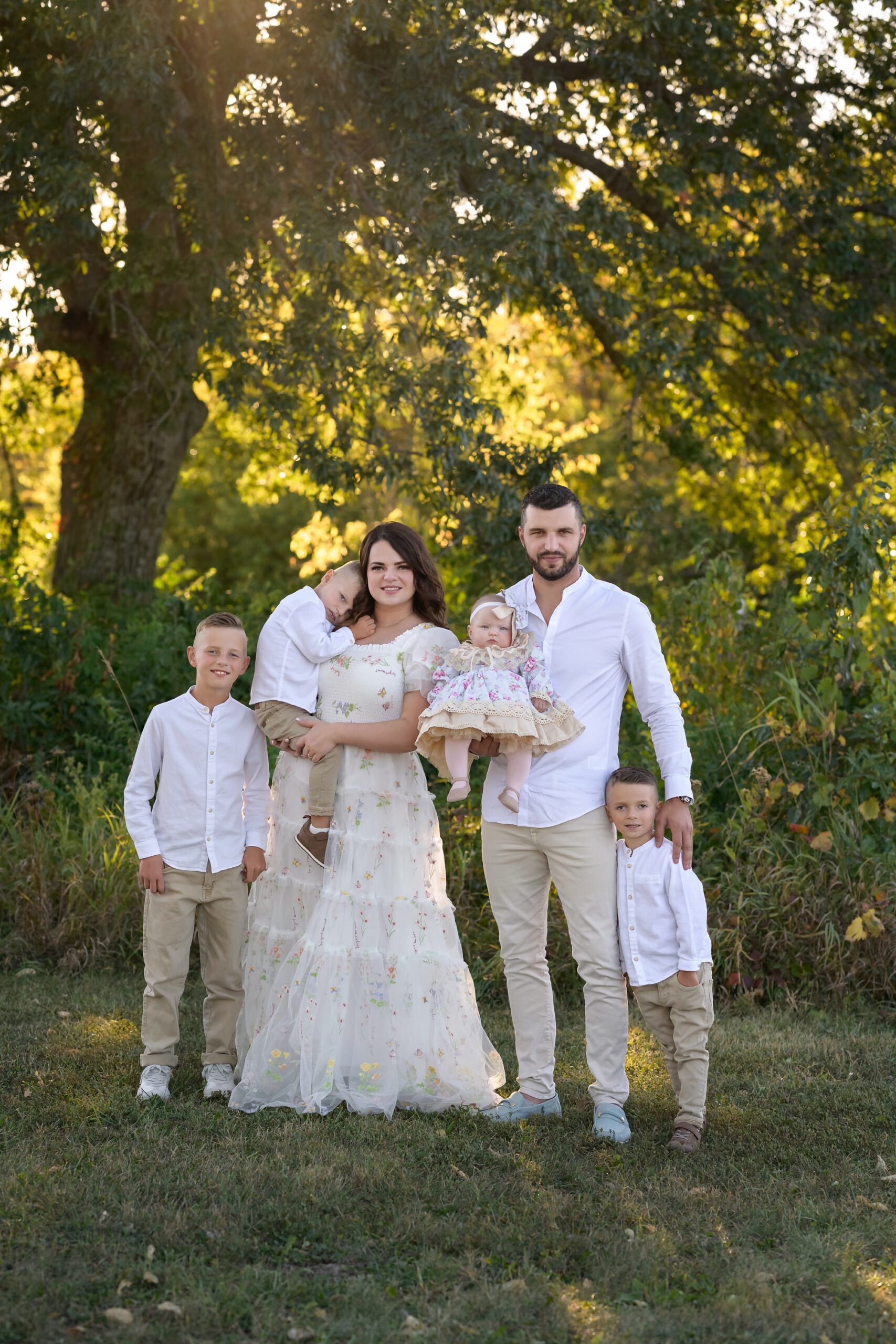 Family of six standing outside, in front of some backlit trees. Family is dressed up in whites, creams, and florals. Photographed in south Kansas City by Helen Ransom.