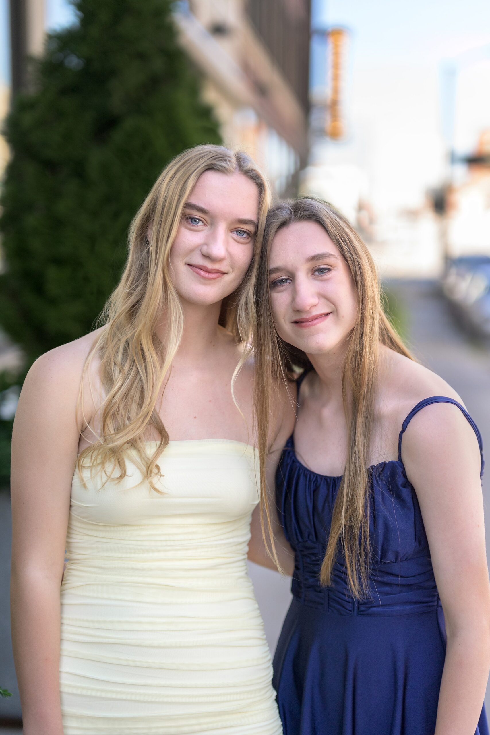 Twin teen girls in yellow and blue dresses, standing with heads together at their picture session in Kansas City.