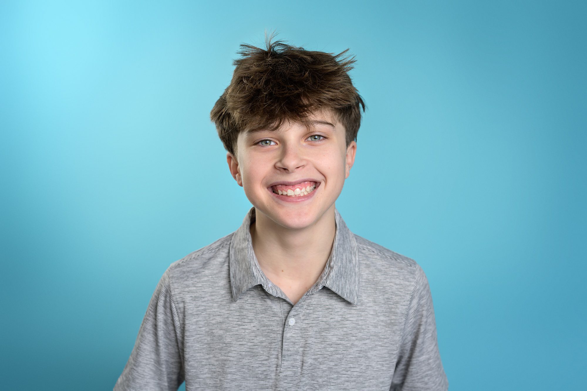 Young teen boy in a gray striped polo standing in front of a bright blue studio background, during a family pictures session in Overland Park.