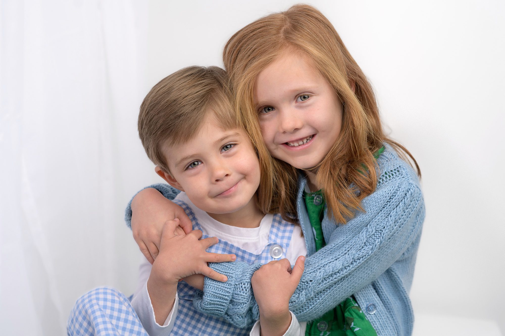 Close-up picture of a brother and sister hugging and looking at the camera. Both are wearing blues, whites, and greens, and were photographed in a white set at Helen Ransom's Kansas City photography studio.