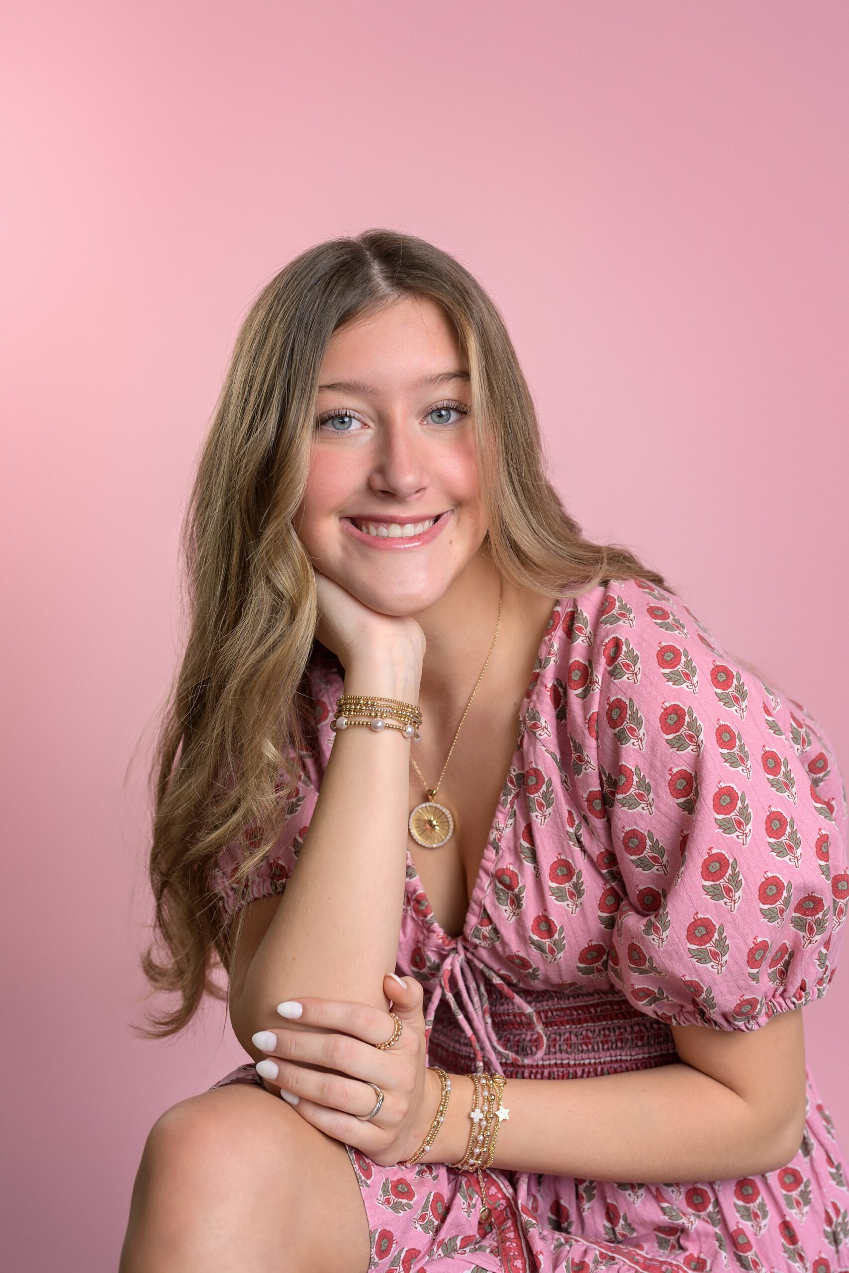 Teen girl with long, light brown hair, wearing a pink floral print dress and posing in front of a pink backdrop at a Kansas City photo session.