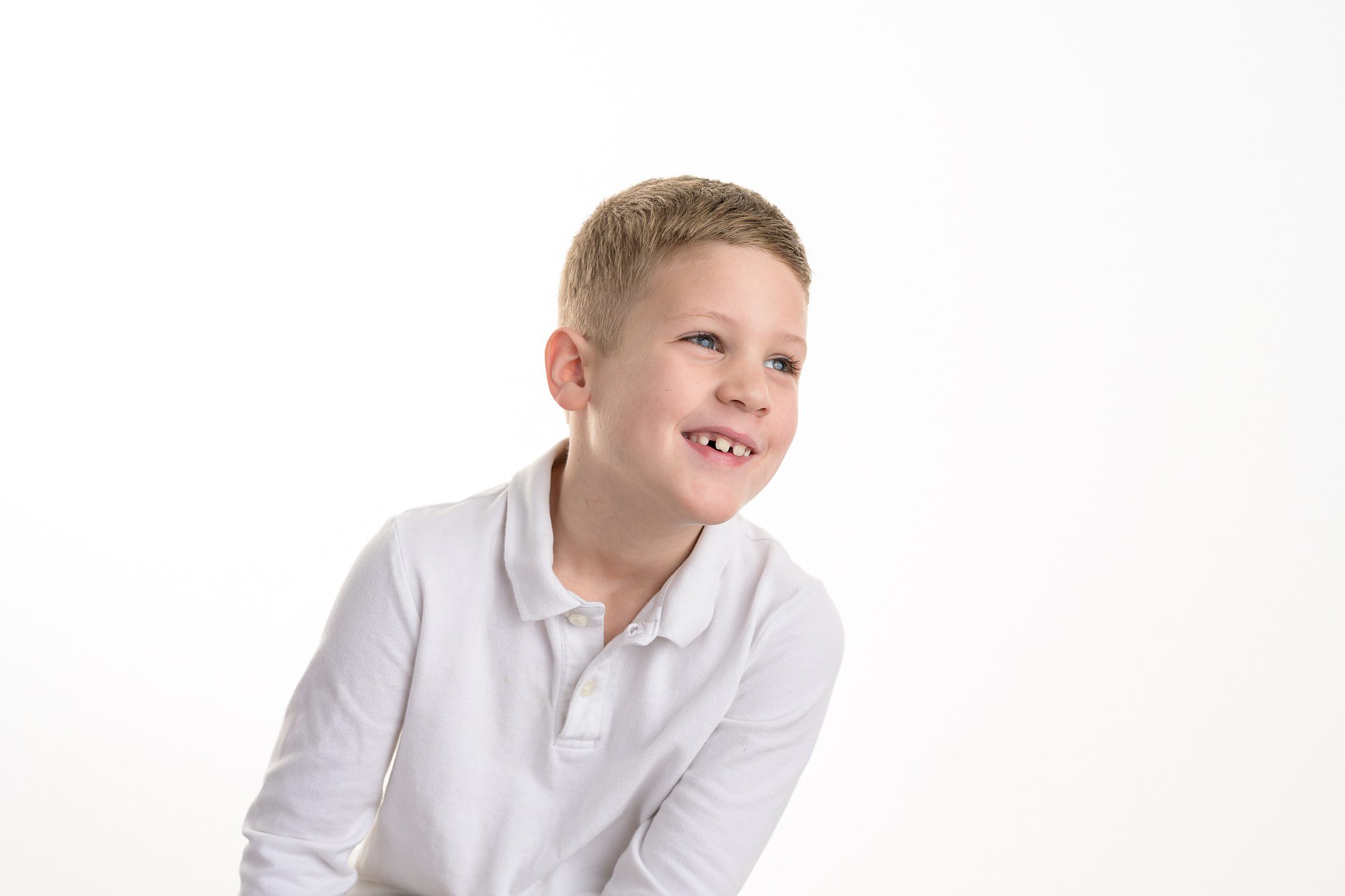 7 year old boy in a white polo, sitting against a white background, leaning and looking off to the right while laughing.