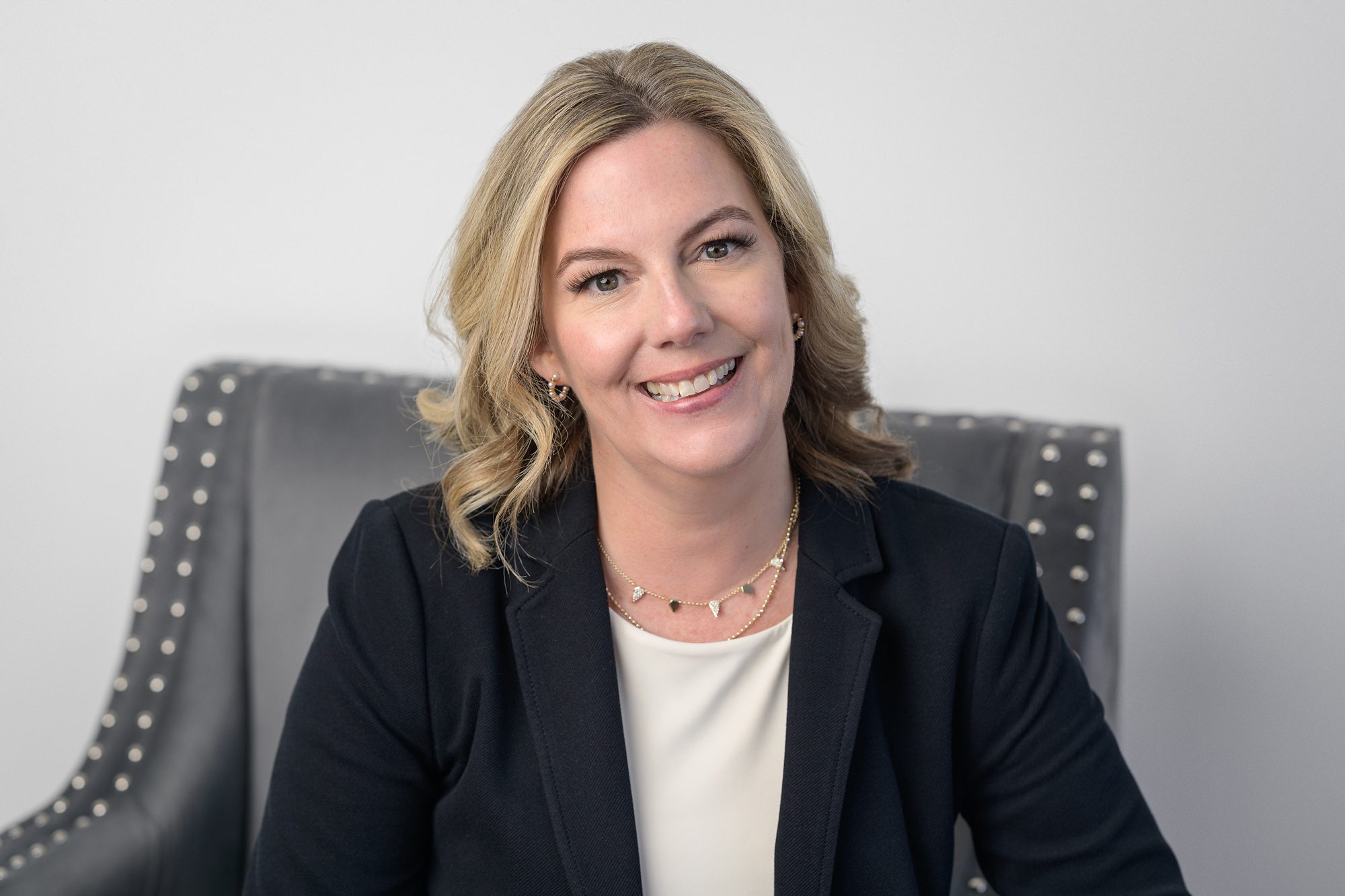 Professional female with shoulder length blonde hair, wearing a black blazer over a white top, and she's sitting in a gray chair for her headshot made in Kansas City.