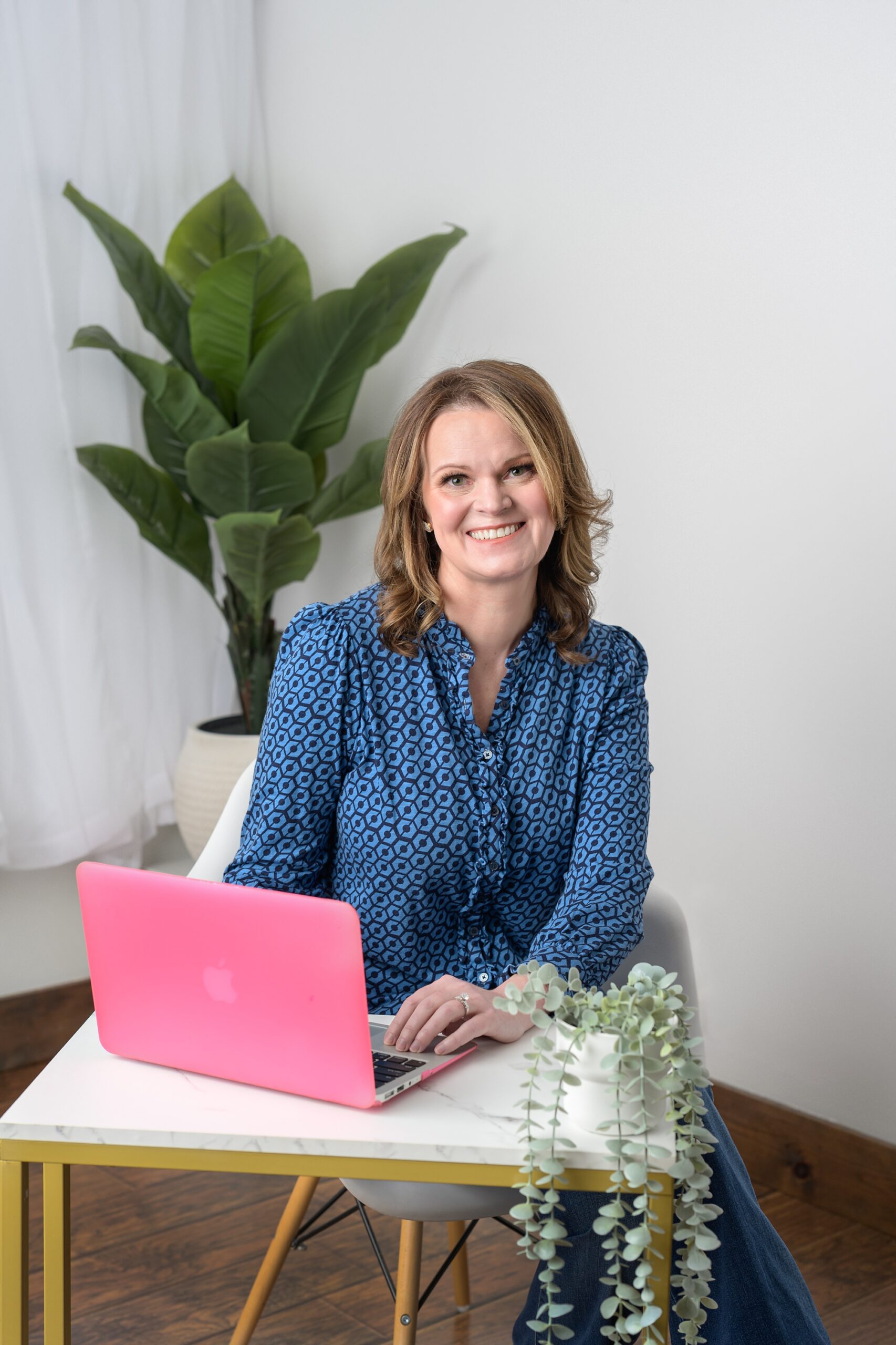 Woman in blue sitting at her hot pink laptop, with hands on the keyboards and she's smiling at the camera, during her personal branding session in Kansas City.