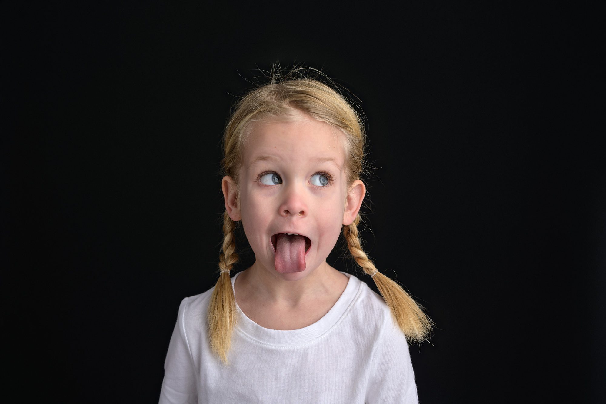 Close up portrait of a 4 year old girl in a white shirt, braids, and sitting in front of a black background at a photo studio in Overland Park. She's looking to the right and sticking her tongue out for a silly picture.
