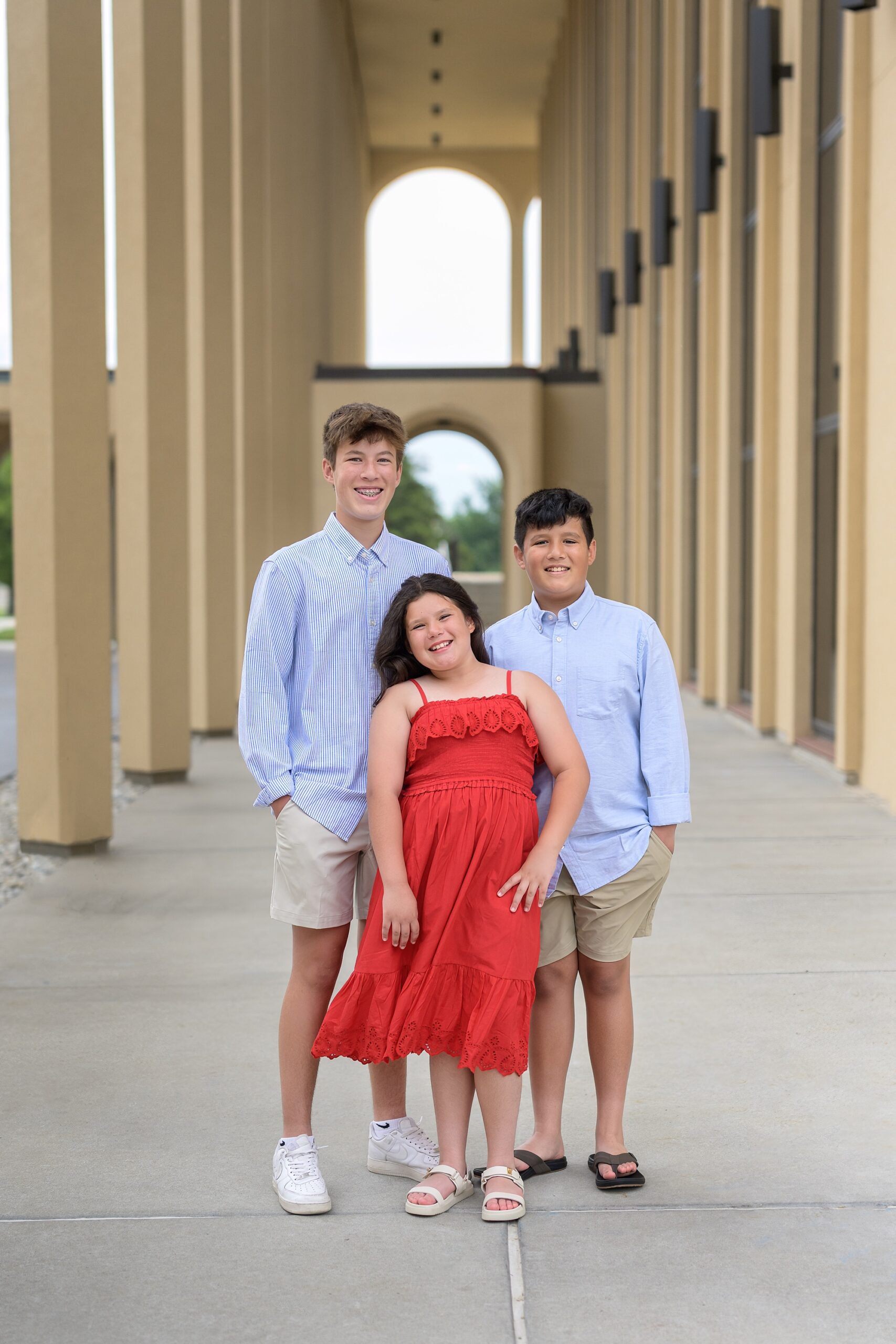 Three siblings, two boys and a girl, standing in a walkway surrounded by large columns. The boys are in button down light blue shirts and the girl is in a red dress.