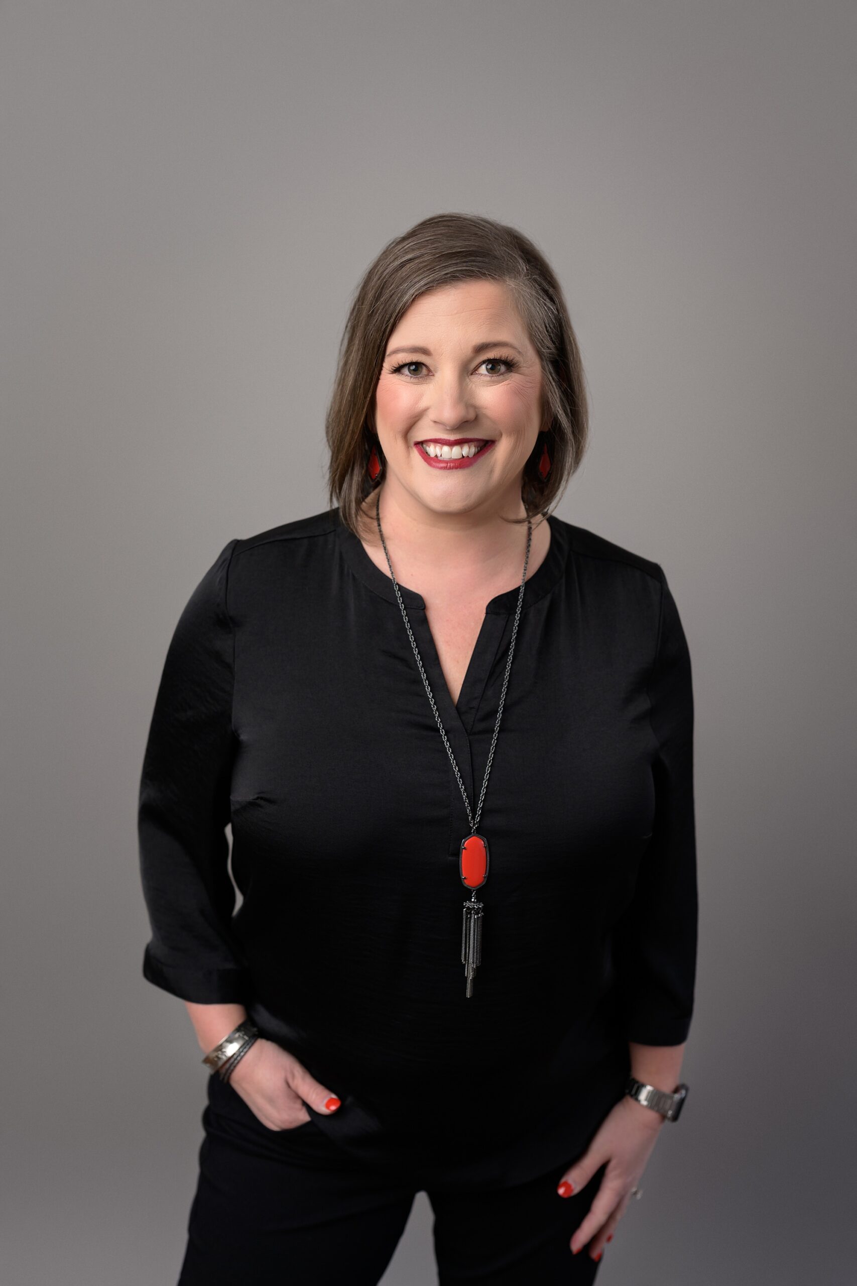 Woman dressed in black, with a bright red pendant and lipstick to match, standing in front of a gray studio backdrop for a more casual take on a headshot.