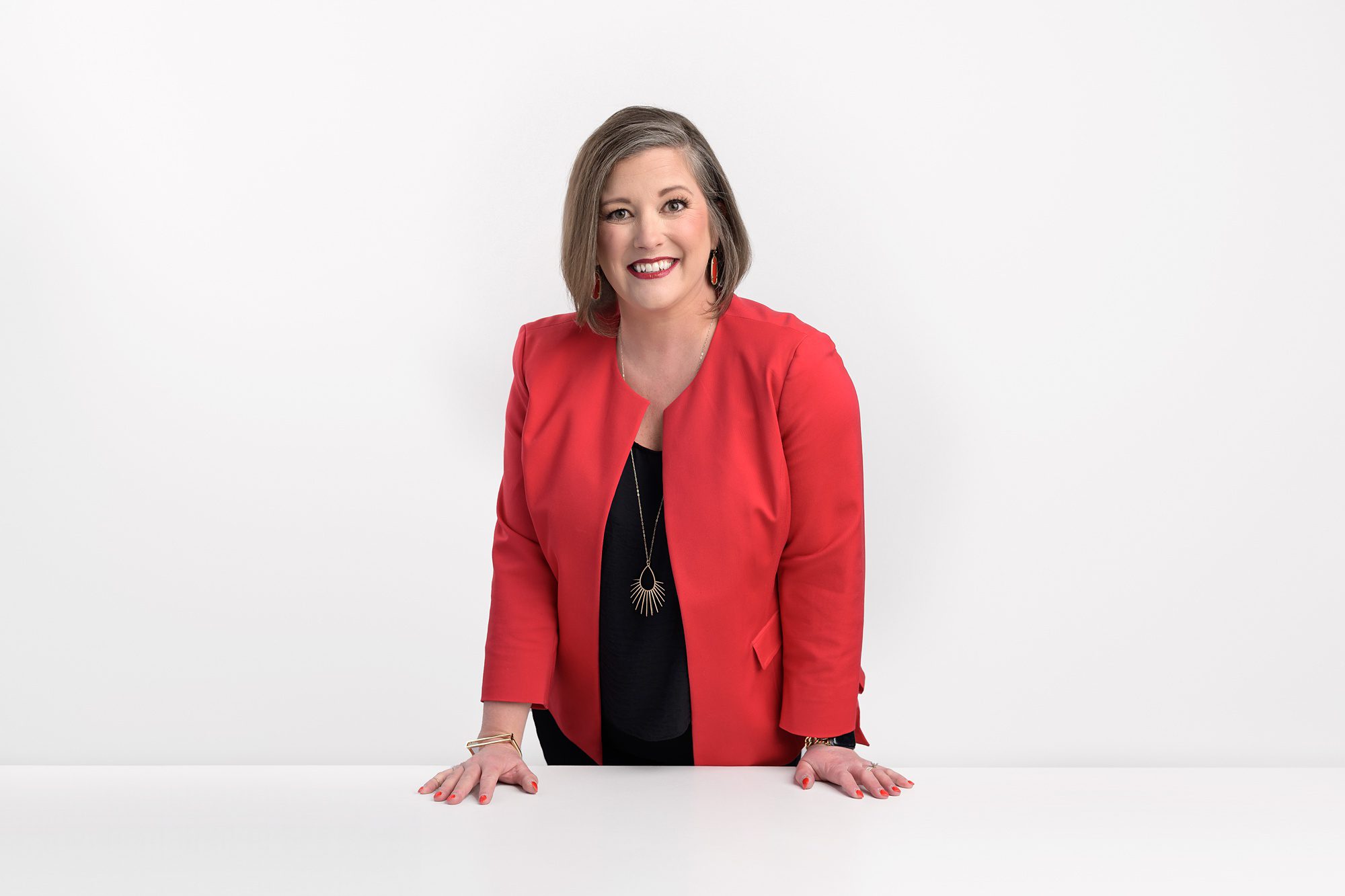 Woman in a red blazer, standing at a white table against a white wall, during her headshot session at a photography studio in Overland Park.