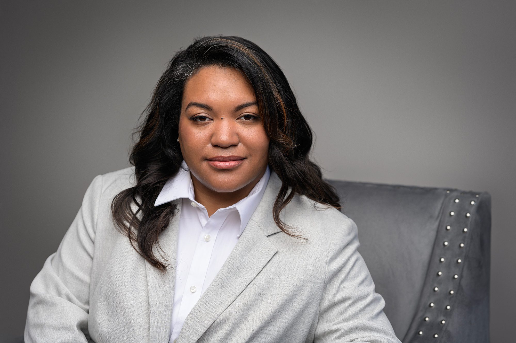 African American business woman, sitting in a chair against a gray studio backdrop in Overland Park. She's staring directly into the camera with a serious, yet pleasant look on her face.