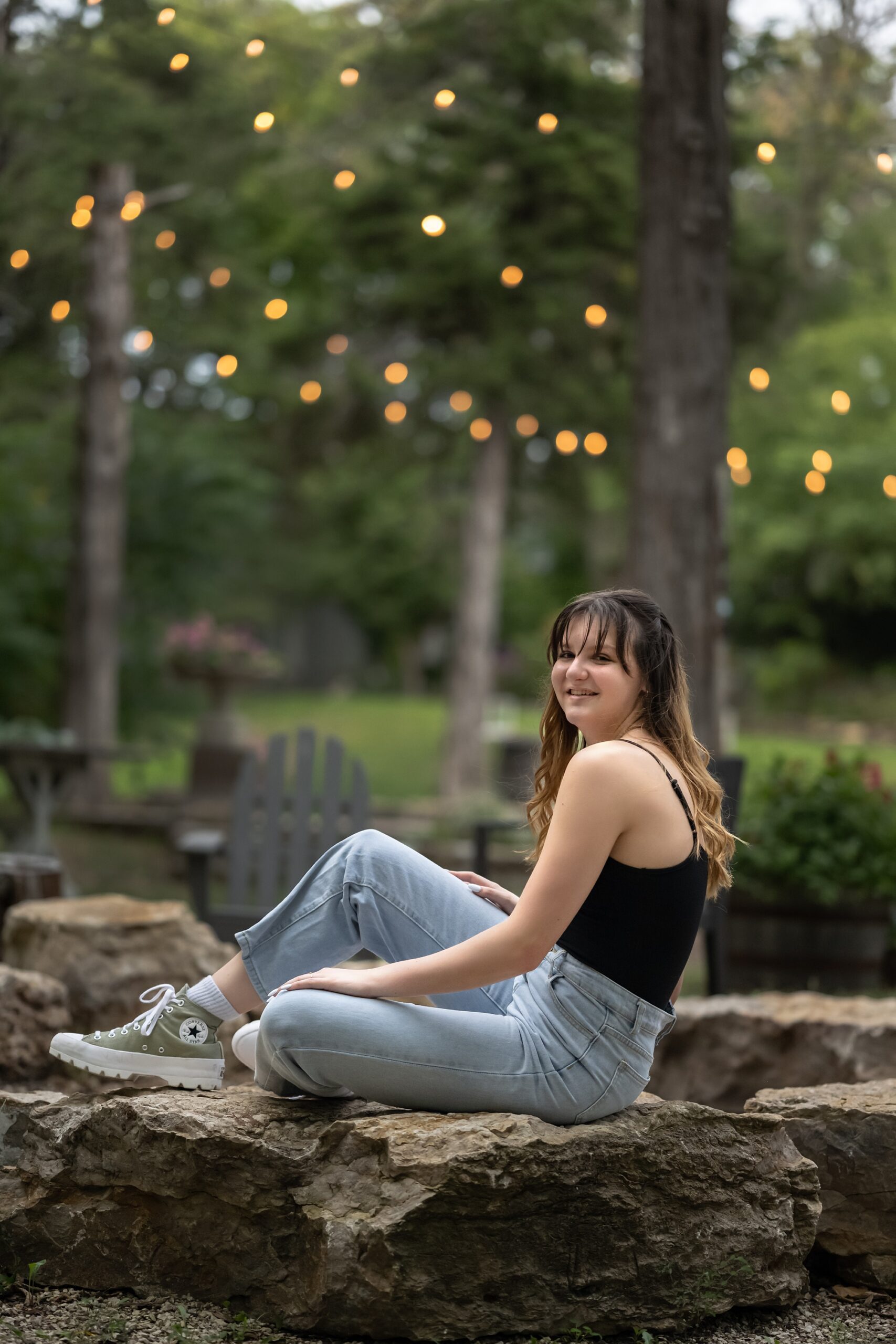 Teen girl in jeans and a black tank top, sitting on large rocks with trees and lights strung behind her, at her senior photo session in Overland Park.