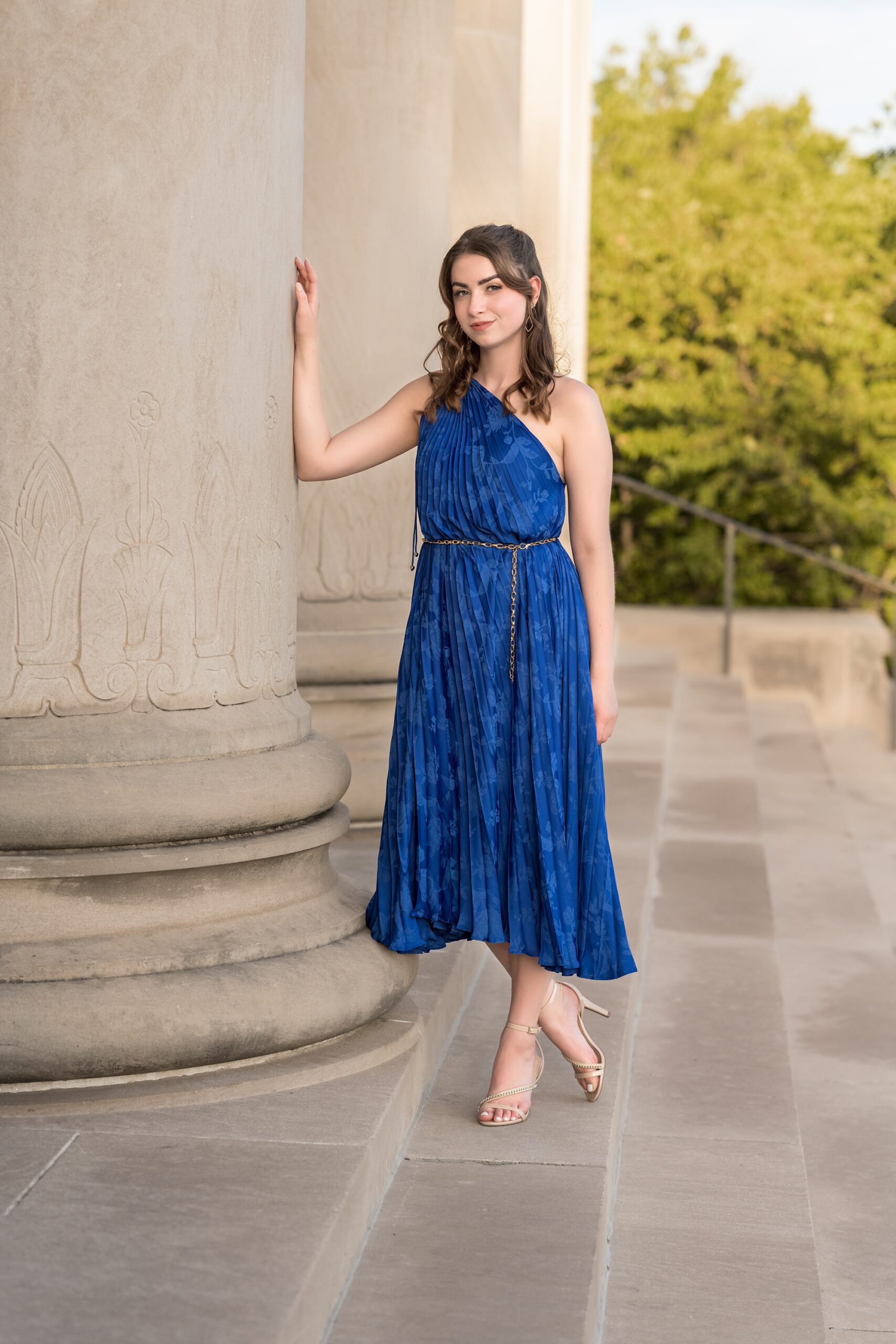 Senior girl in a long, royal blue dress, leaning against one of the columns at the Nelson Atkins Museum of Art in Kansas City.