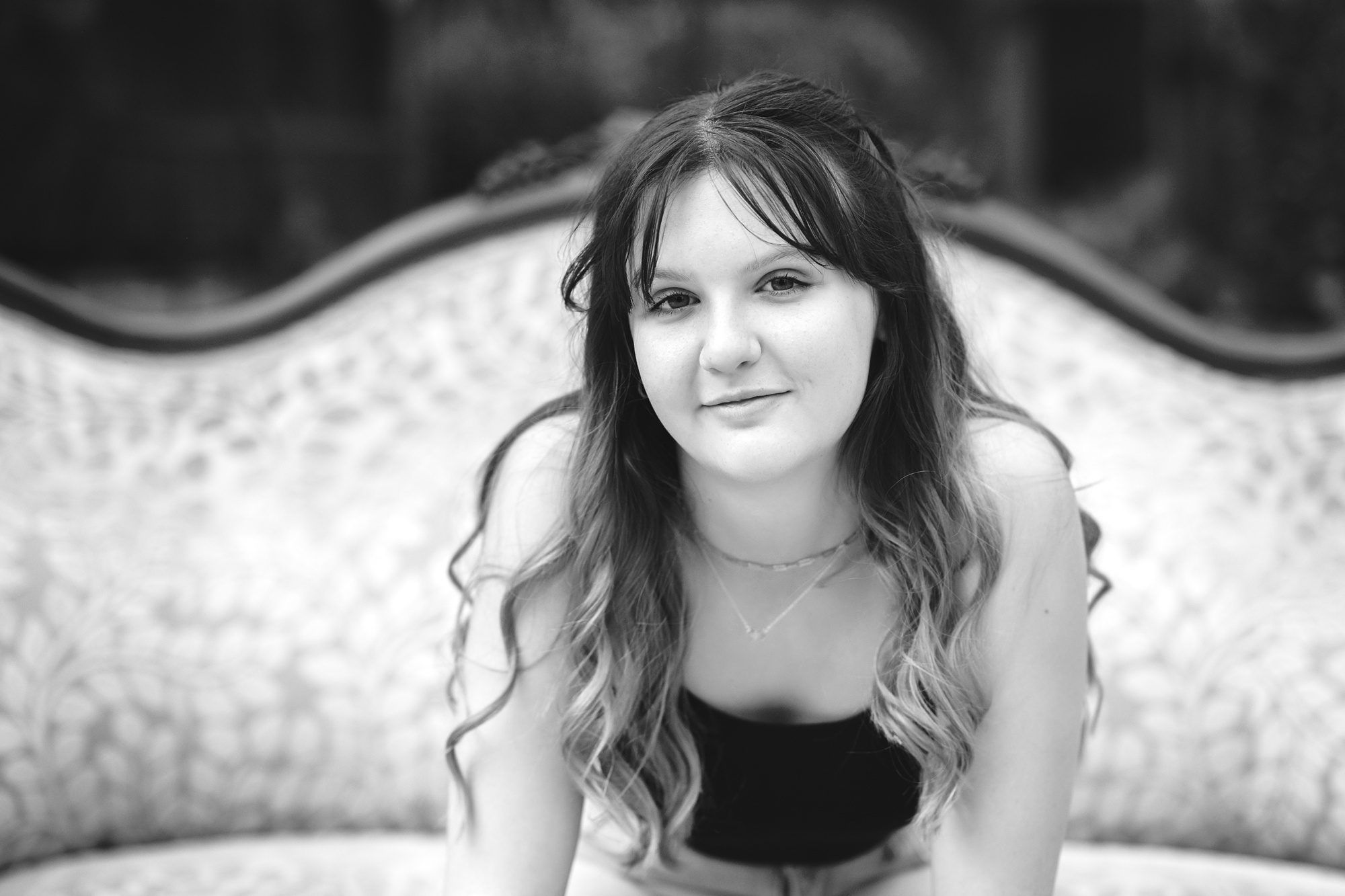 Black and white close up of a teen girl sitting on a couch, leaning forward, and looking at the camera with a serious expression.