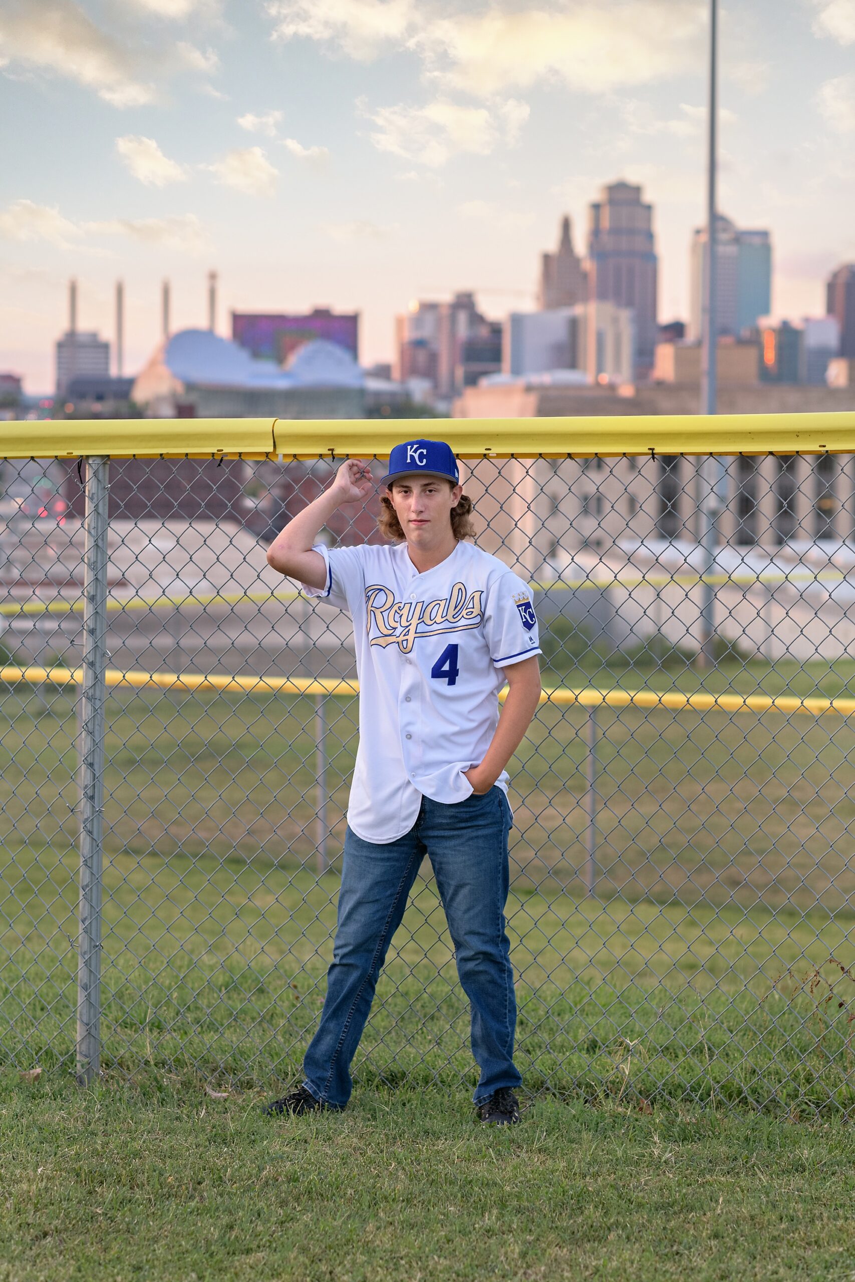 High school guy standing against an outfield fence, with the Kansas City skyline behind him. He's wearing his favorite Royals jersey and hat, with jeans.