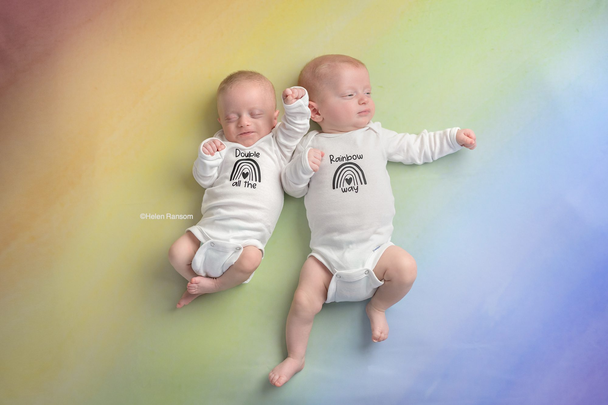 Twin baby boys on a rainbow background, wearing onesies calling them double rainbow blessings. These babies are known as rainbow babies, because they were born after a loss.
